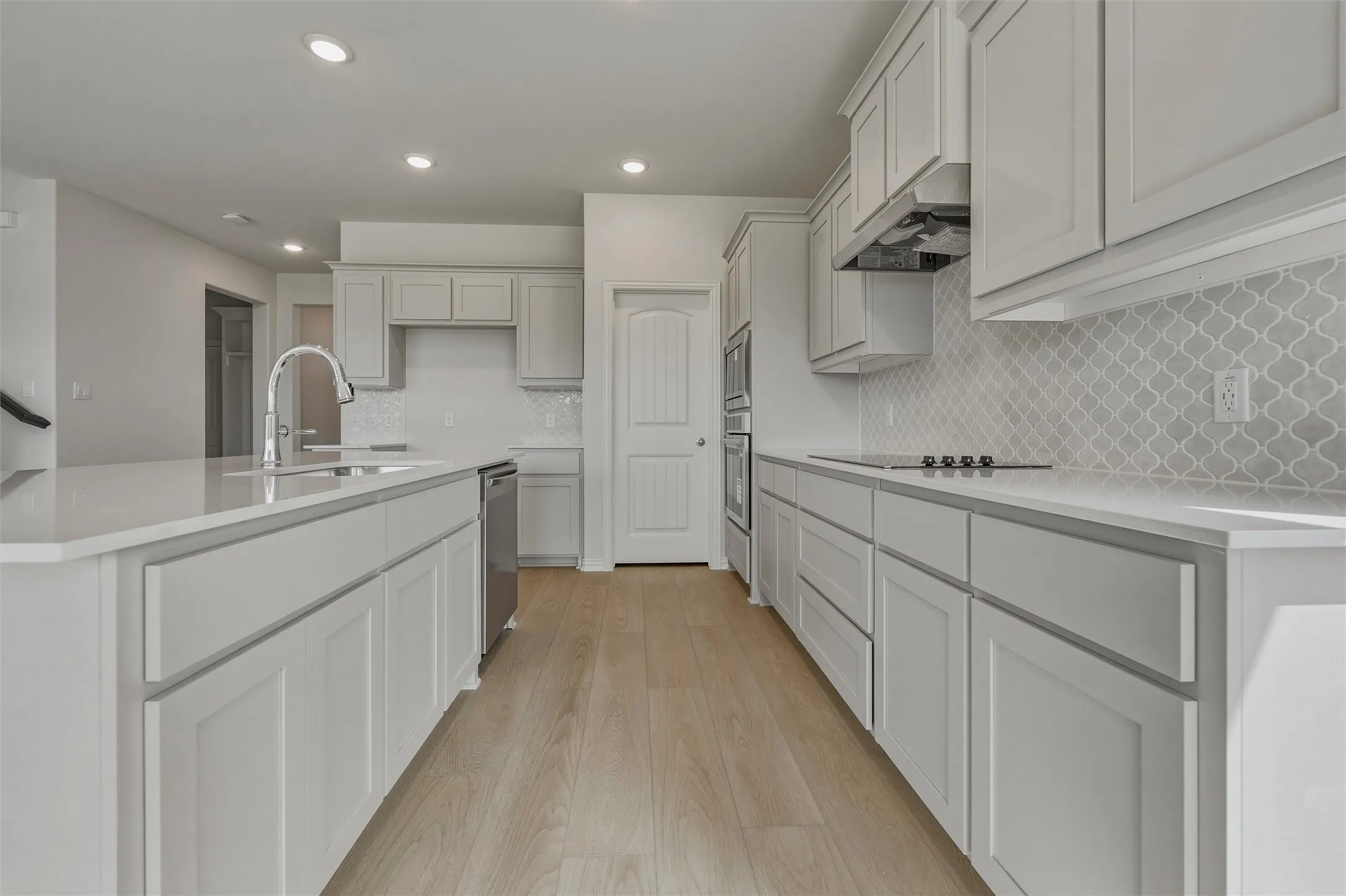 Kitchen featuring light-toned cabinetry, an expansive island with a stainless steel sink, light-colored countertops, a decorative tile backsplash, and wood-finish flooring
