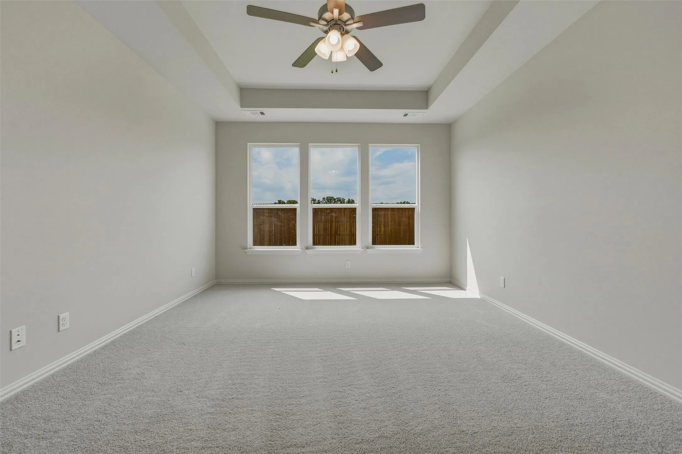 Tray ceiling with a ceiling fan, light gray carpet, and three windows offering natural illumination