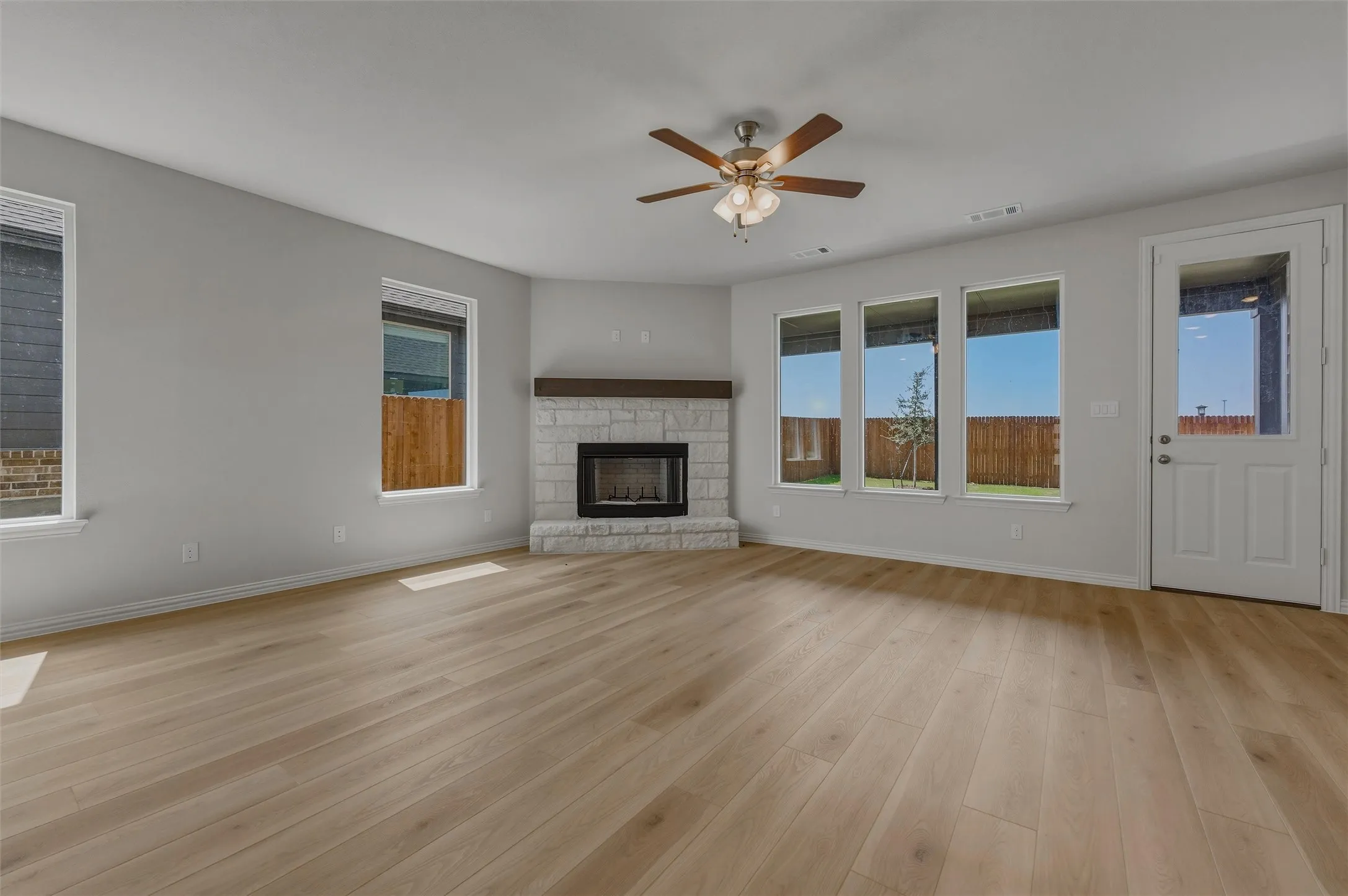 Wood-finish flooring, stone-clad fireplace with wood mantle, multiple windows, and a ceiling fan