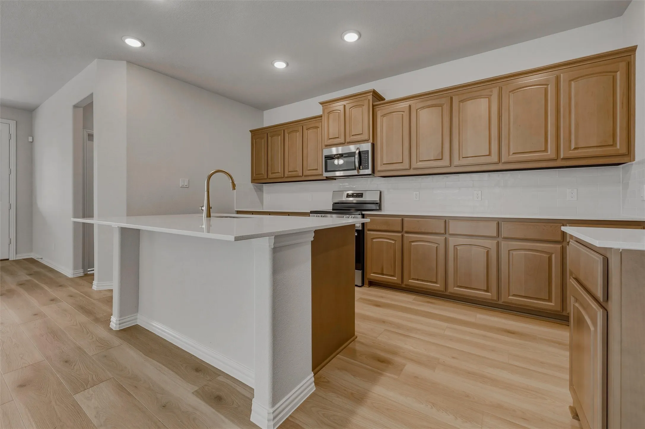Kitchen featuring wood-finish flooring, recessed lighting, a kitchen island with a white countertop and brass-finish faucet, extensive cabinetry, and stainless steel appliances
