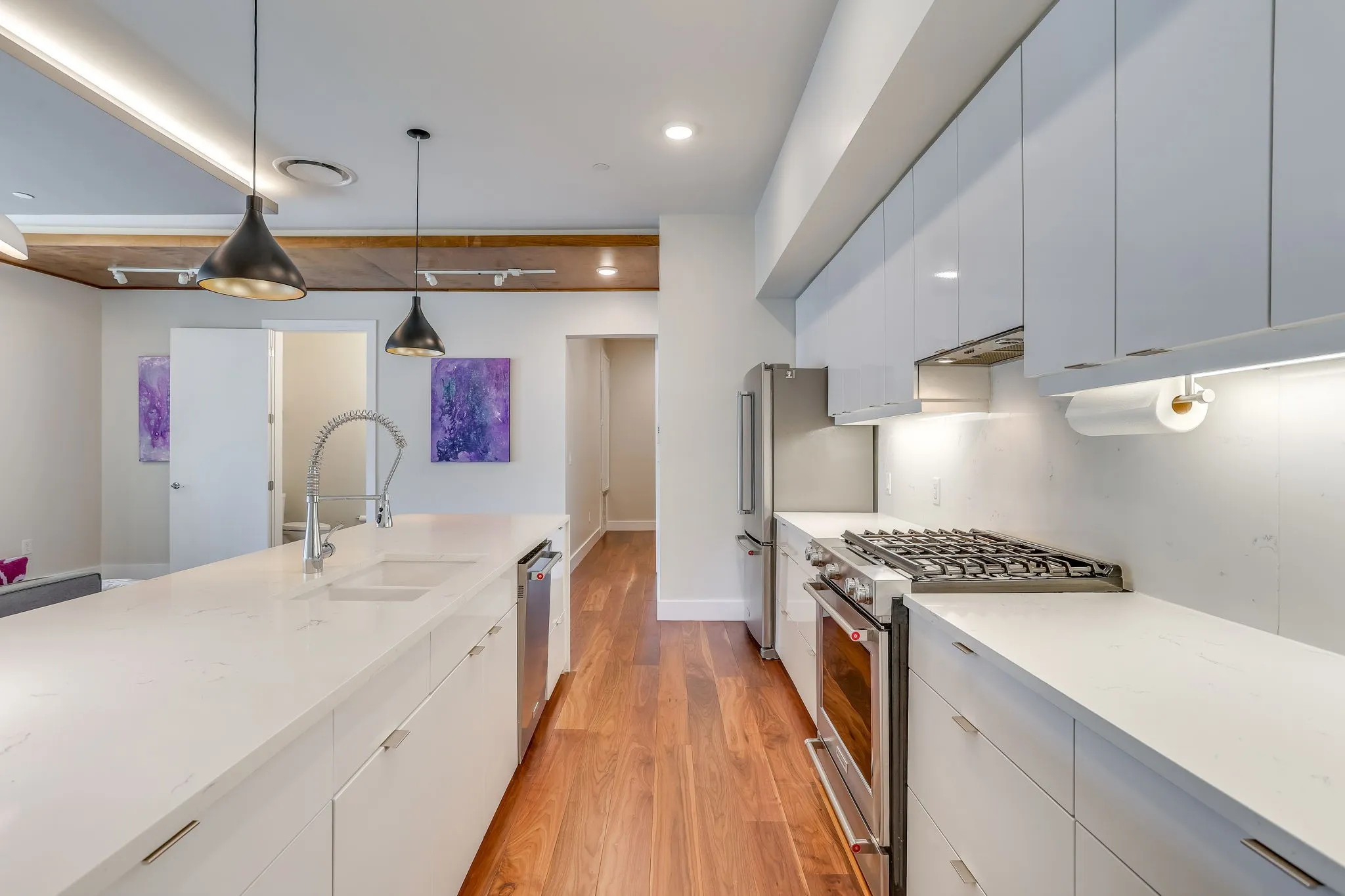 Modern kitchen featuring an expansive island with an undermount sink, white gloss cabinetry, stainless steel appliances, Carlisle wide plank wood flooring, and pendant lighting
