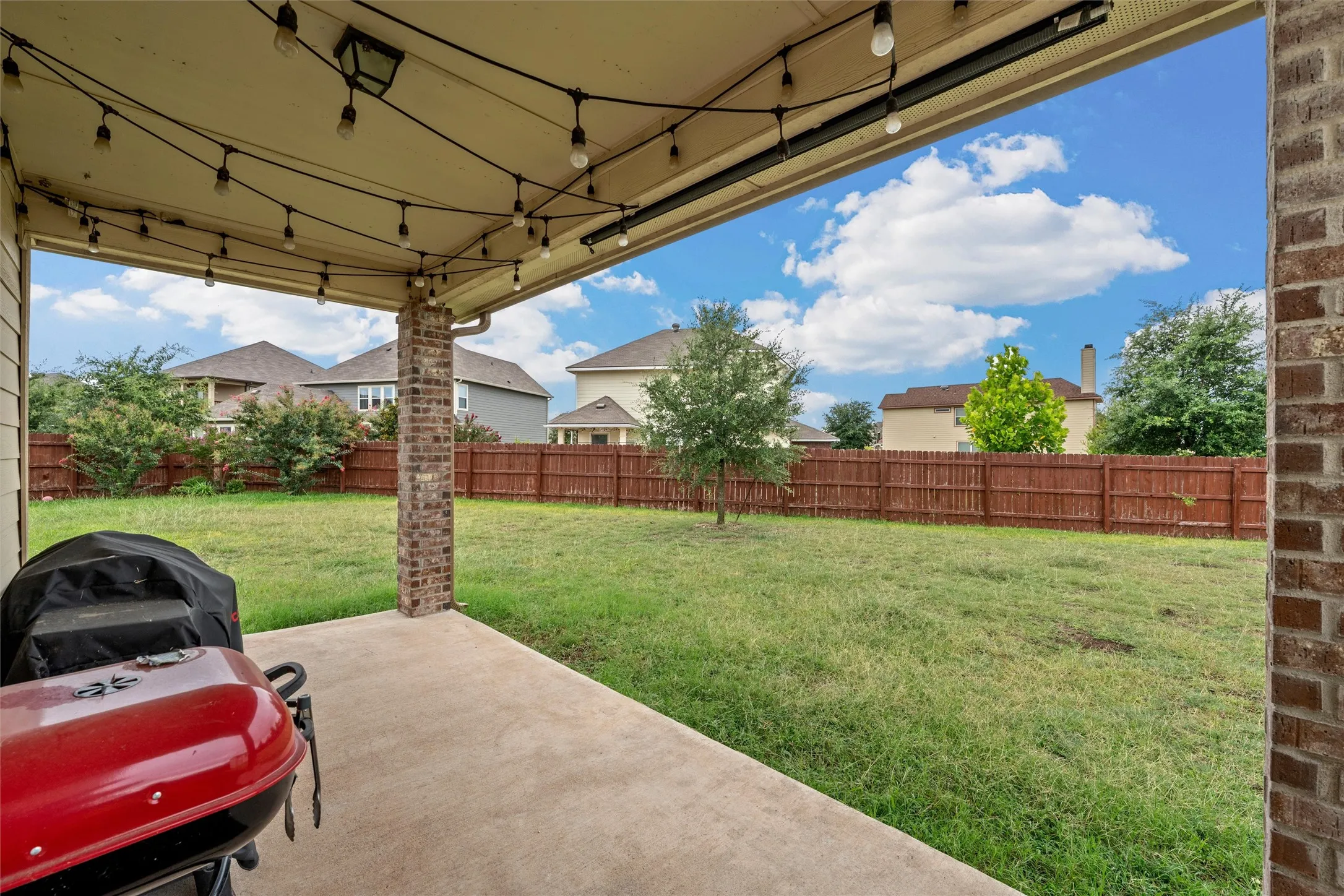 Covered concrete patio with brick support columns, string lighting, and a spacious backyard featuring a wood privacy fence and mature landscaping