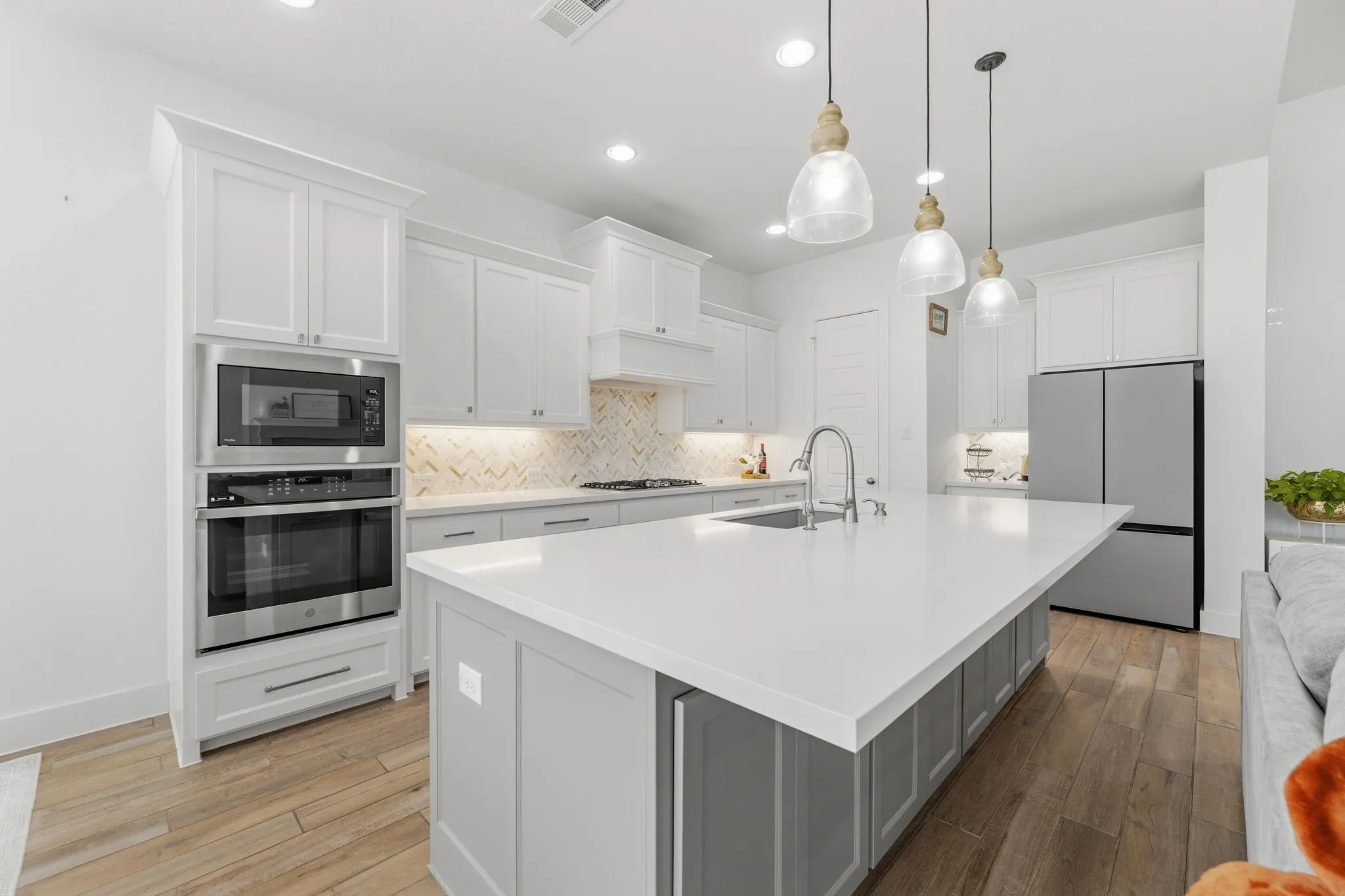 Modern kitchen featuring white cabinetry, a built-in stainless steel microwave and oven, a chevron tile backsplash, a large island with a sink, and wood-finish flooring