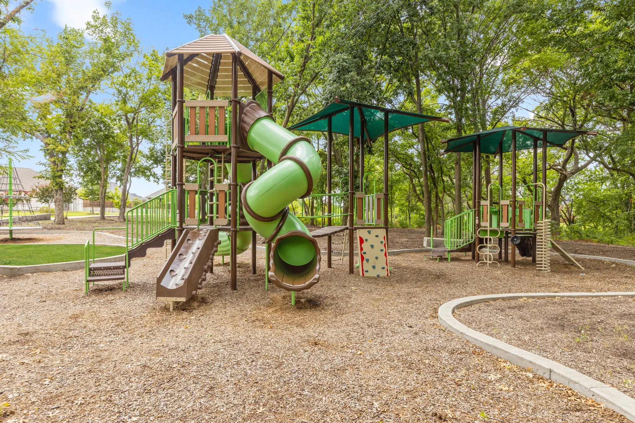 Outdoor recreational area featuring a multi-level play structure with a spiral slide, climbing wall, and shaded platforms