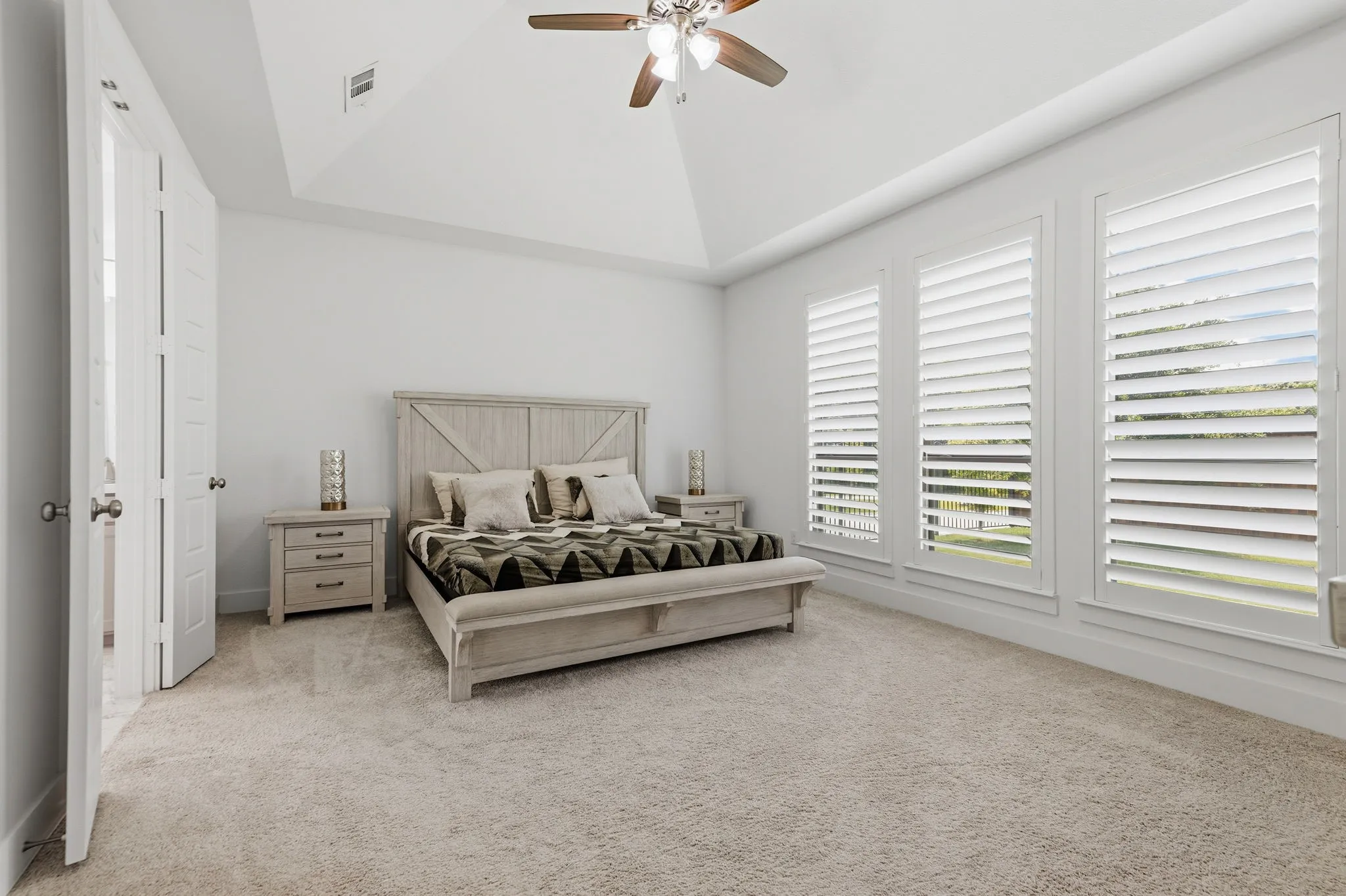 Spacious room featuring vaulted ceilings, a ceiling fan, plush carpeting, three large windows with plantation shutters, and a paneled interior door