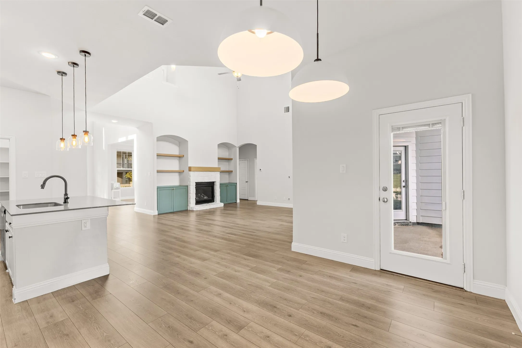 Bright open-concept interior featuring vaulted ceilings, wood-finish flooring, a kitchen island with sink and pendant lighting, and a fireplace with built-in shelving