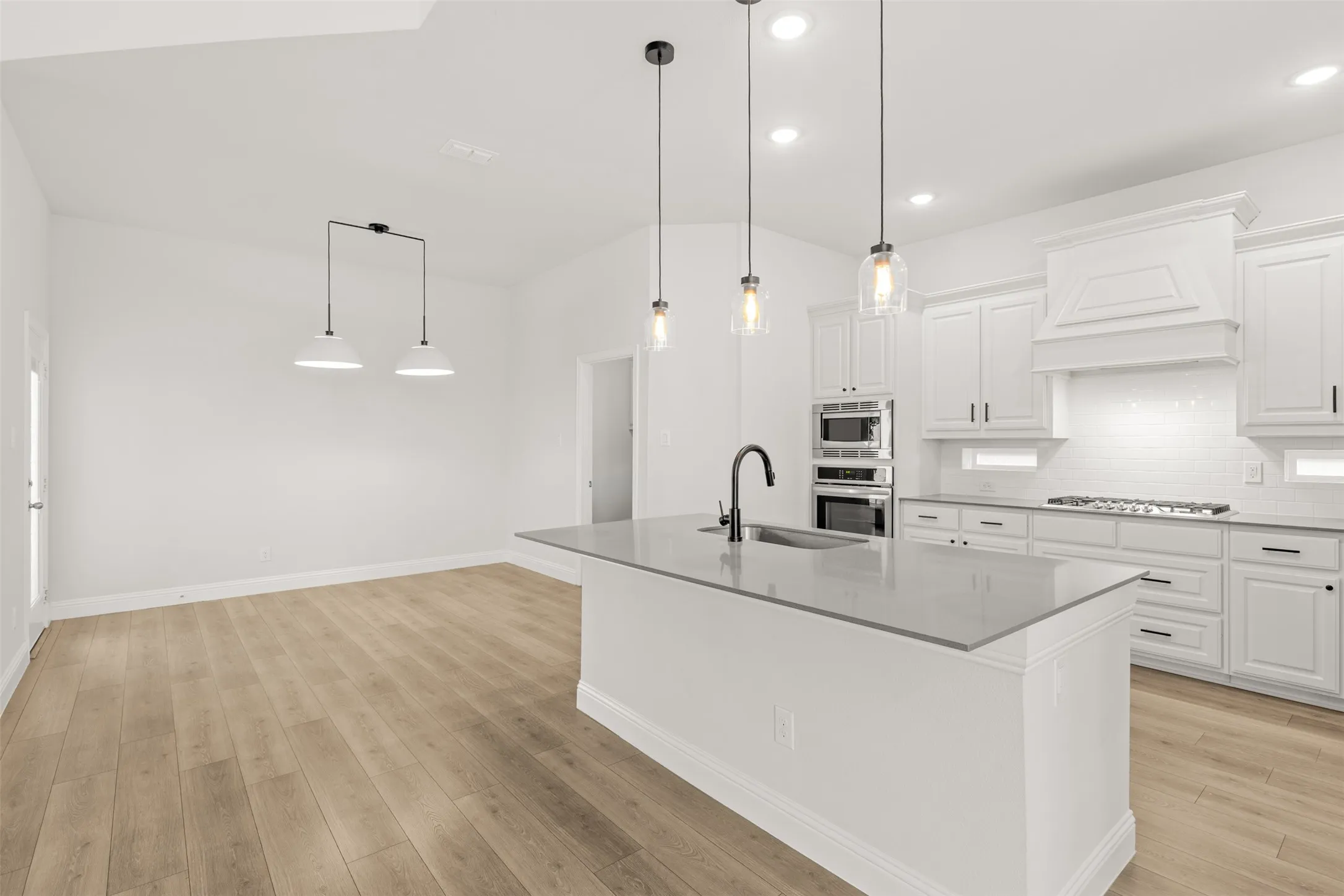 Open-concept kitchen featuring an island with a built-in sink, pendant lighting, white cabinetry, integrated stainless steel appliances, and wood-finish flooring