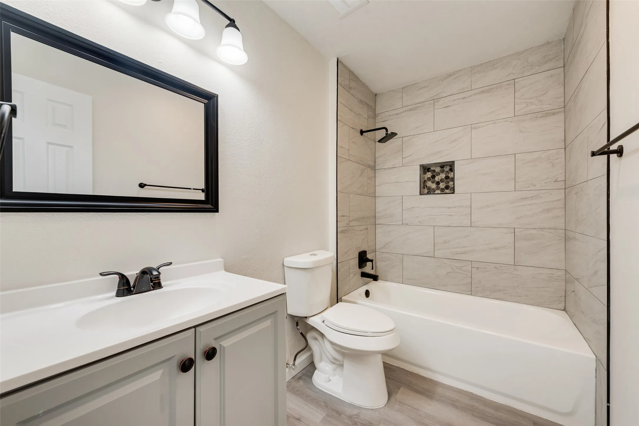 Bathroom featuring a bathtub with light-toned rectangular tile surround, a built-in shower niche with mosaic tile, a rain showerhead, a gray vanity with a white countertop and integrated sink, and wood-finish flooring