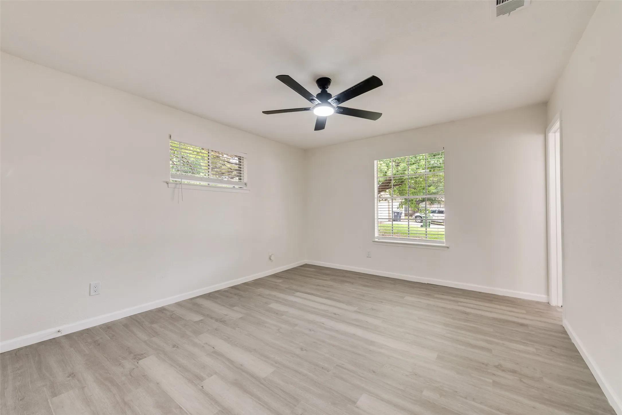 Interior room featuring light-toned wood-finish flooring, two windows with blinds, a ceiling fan with integrated lighting, and white baseboards