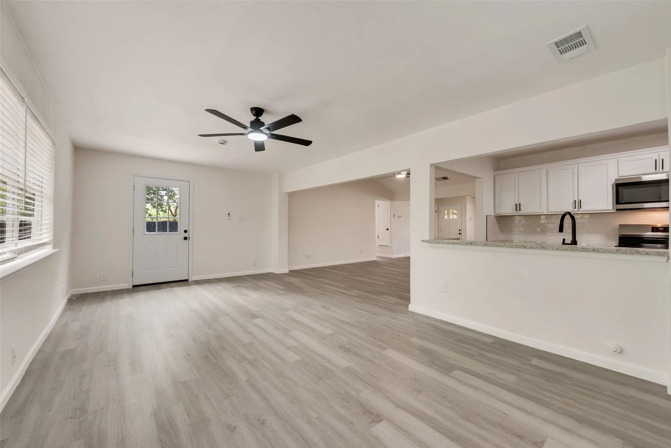 Spacious living area featuring light wood-finish flooring, a contemporary ceiling fan, and a prominent window with horizontal blinds