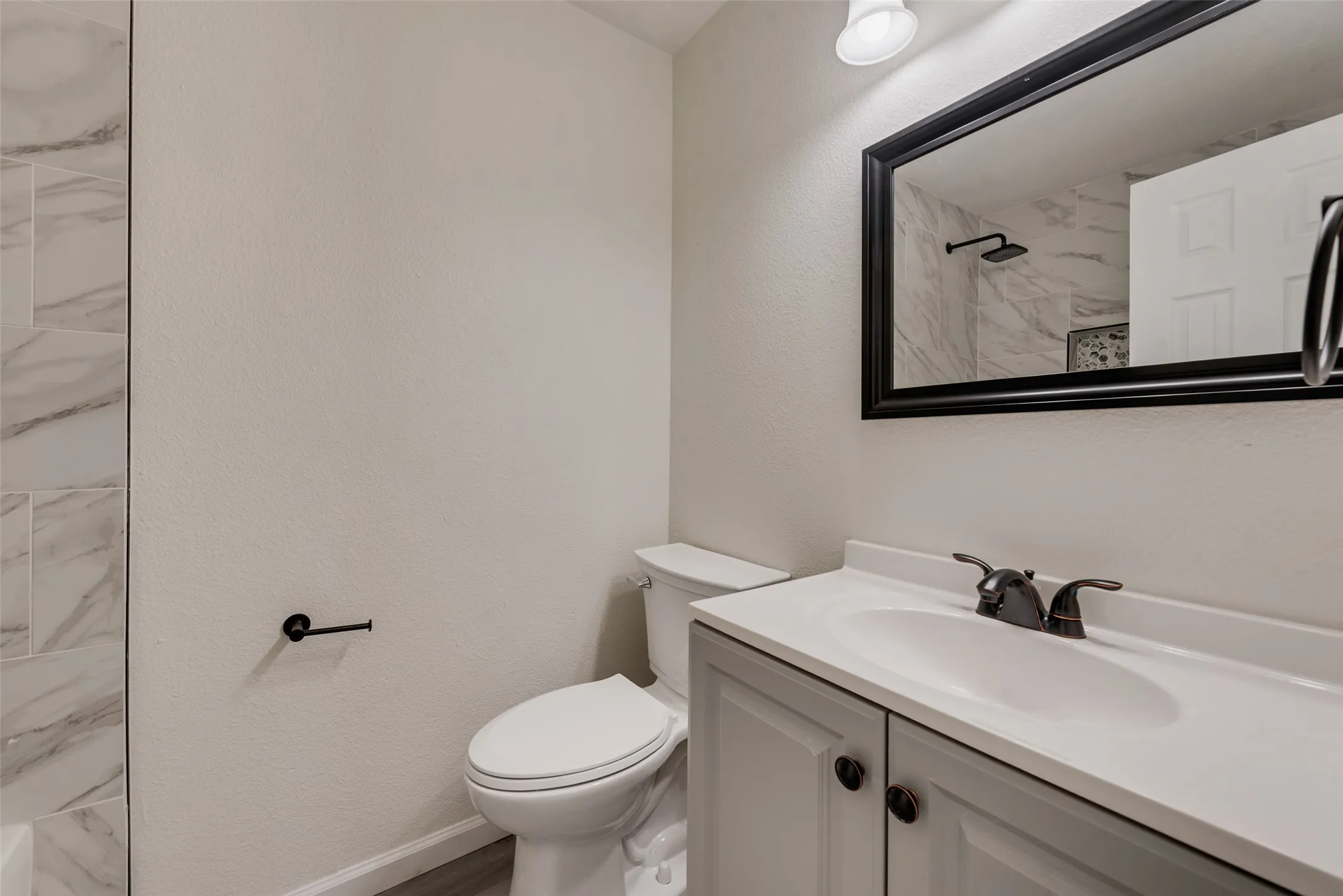 Bathroom featuring a single vanity with a white countertop and gray cabinetry, a rectangular framed mirror, oil-rubbed bronze fixtures, and shower with marble-look tile surround
