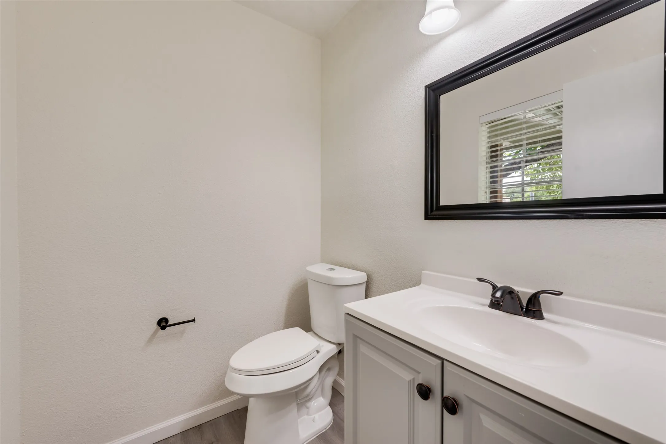 Bathroom featuring light gray walls, wood-finish flooring, a white toilet, and a vanity with a white countertop, gray cabinetry, and dark bronze hardware