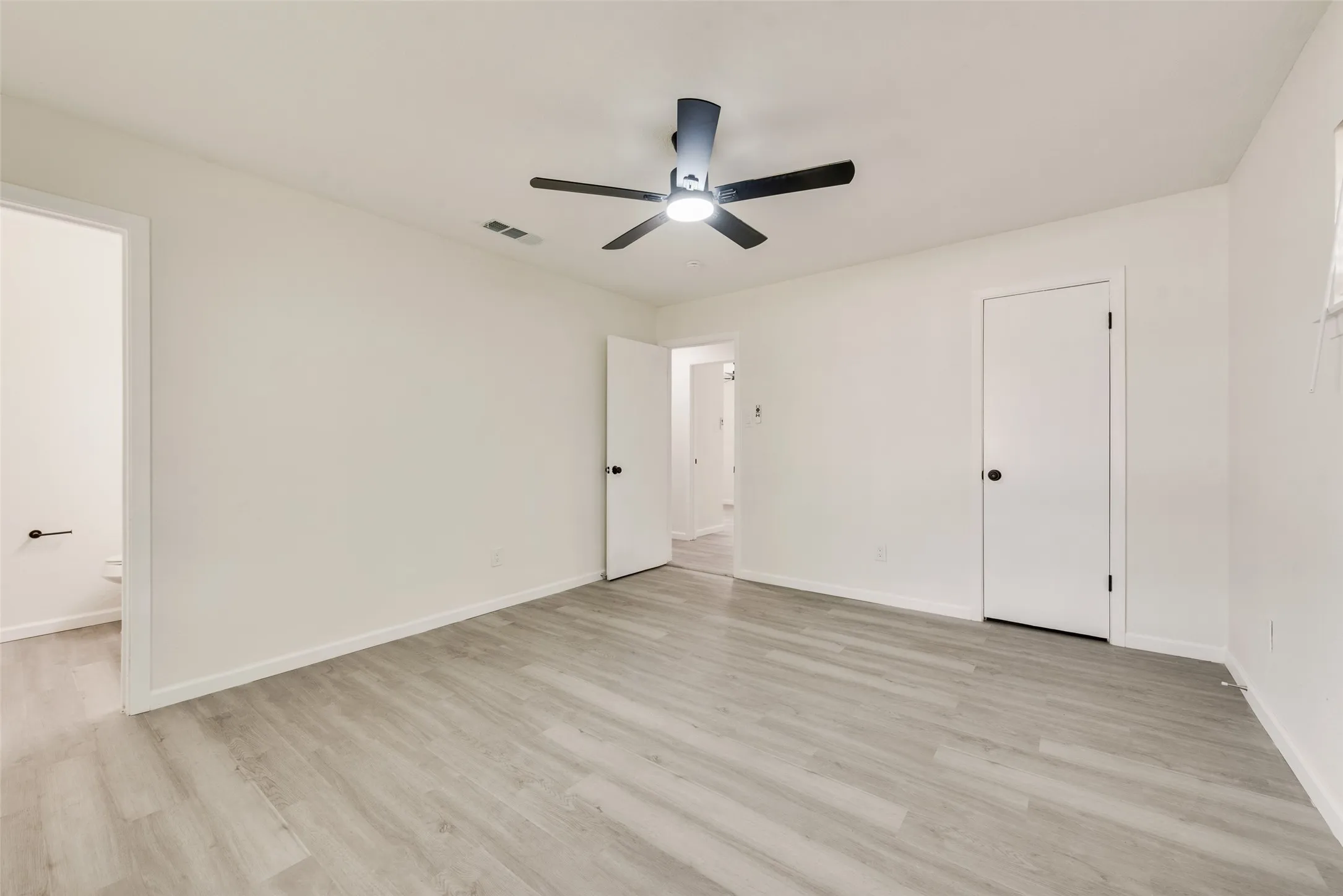 Spacious room featuring light wood-finish flooring, white walls, a ceiling fan with integrated lighting, and multiple doorways