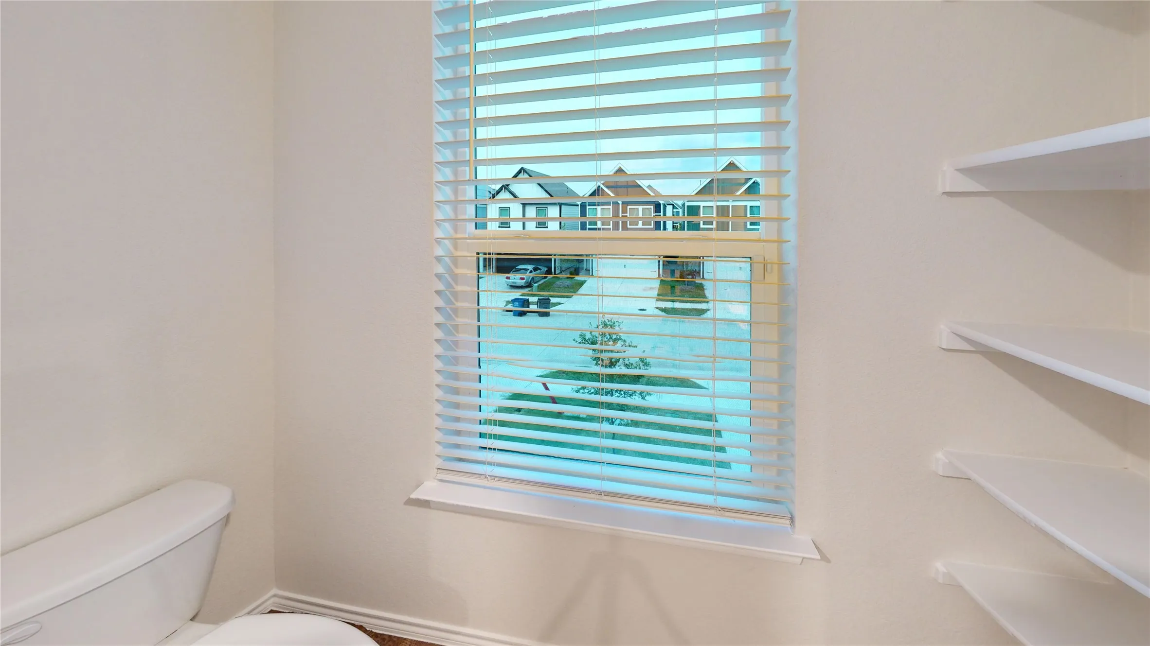 Bathroom window with blinds, white walls, built-in shelving, and a toilet fixture