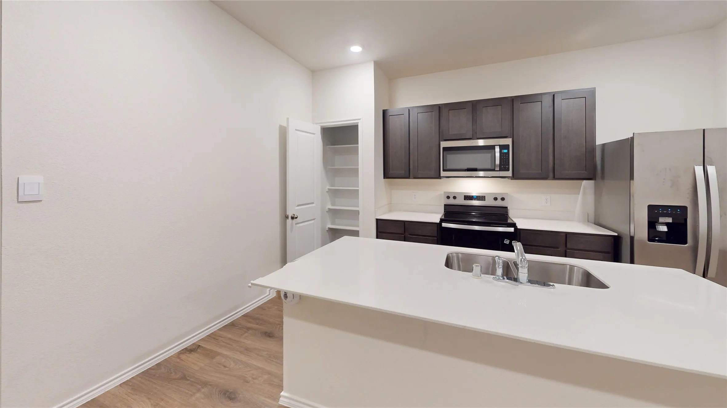 Modern kitchen featuring a large island with an undermount sink, stainless steel appliances, dark wood-finish cabinetry, a white countertop, and wood-finish flooring