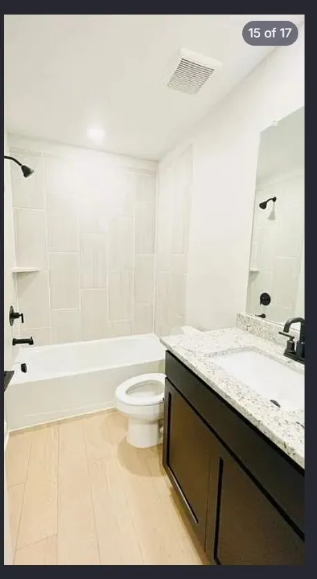Bathroom featuring a white bathtub, light-toned vertical tile surround with an integrated niche, black fixtures, dark wood-finish vanity with a white countertop and integrated sink, and light wood-finish flooring