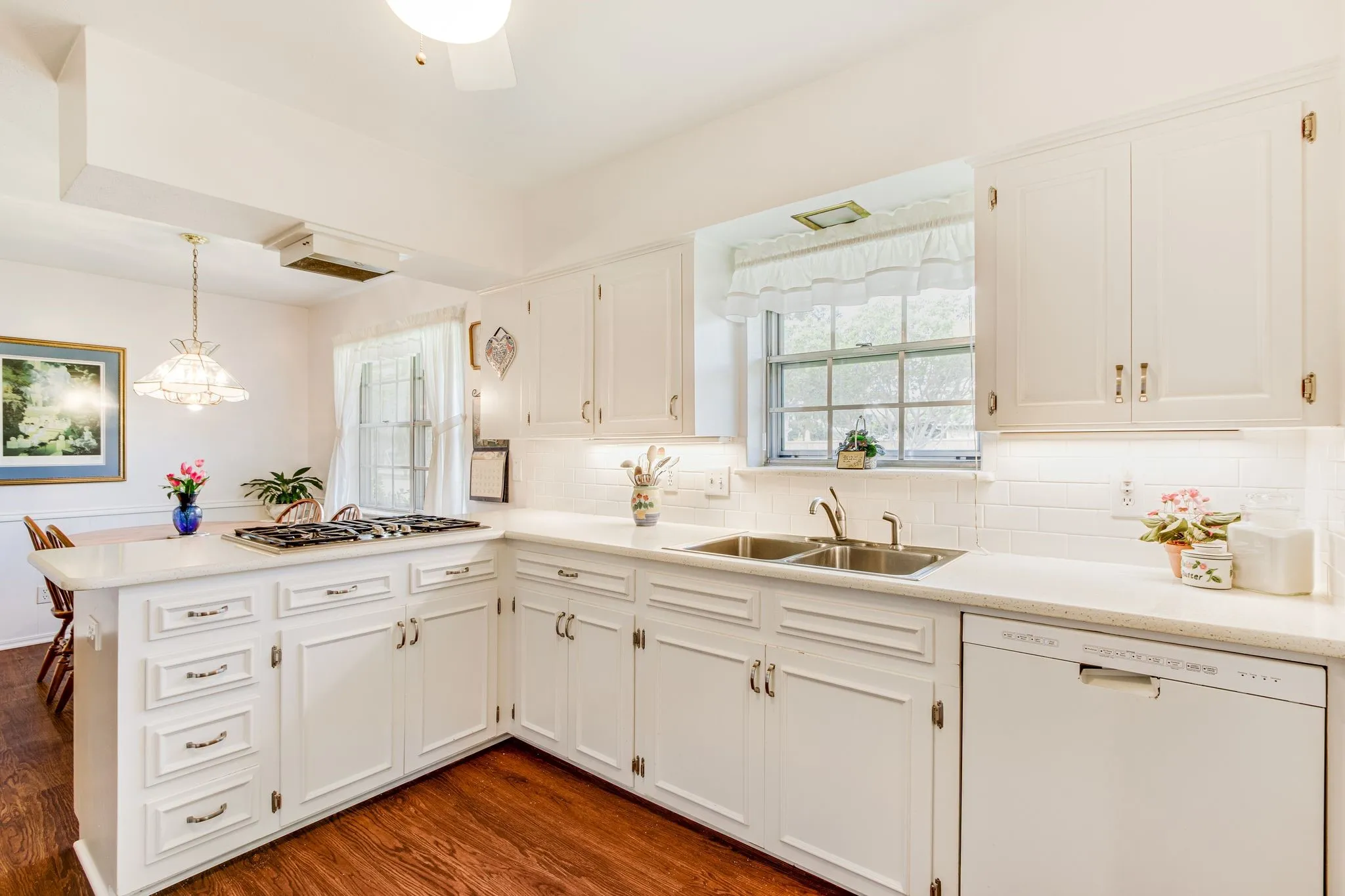 Kitchen featuring white cabinetry, light-toned countertops, a stainless steel double sink, white subway tile backsplash, and wood-finish flooring