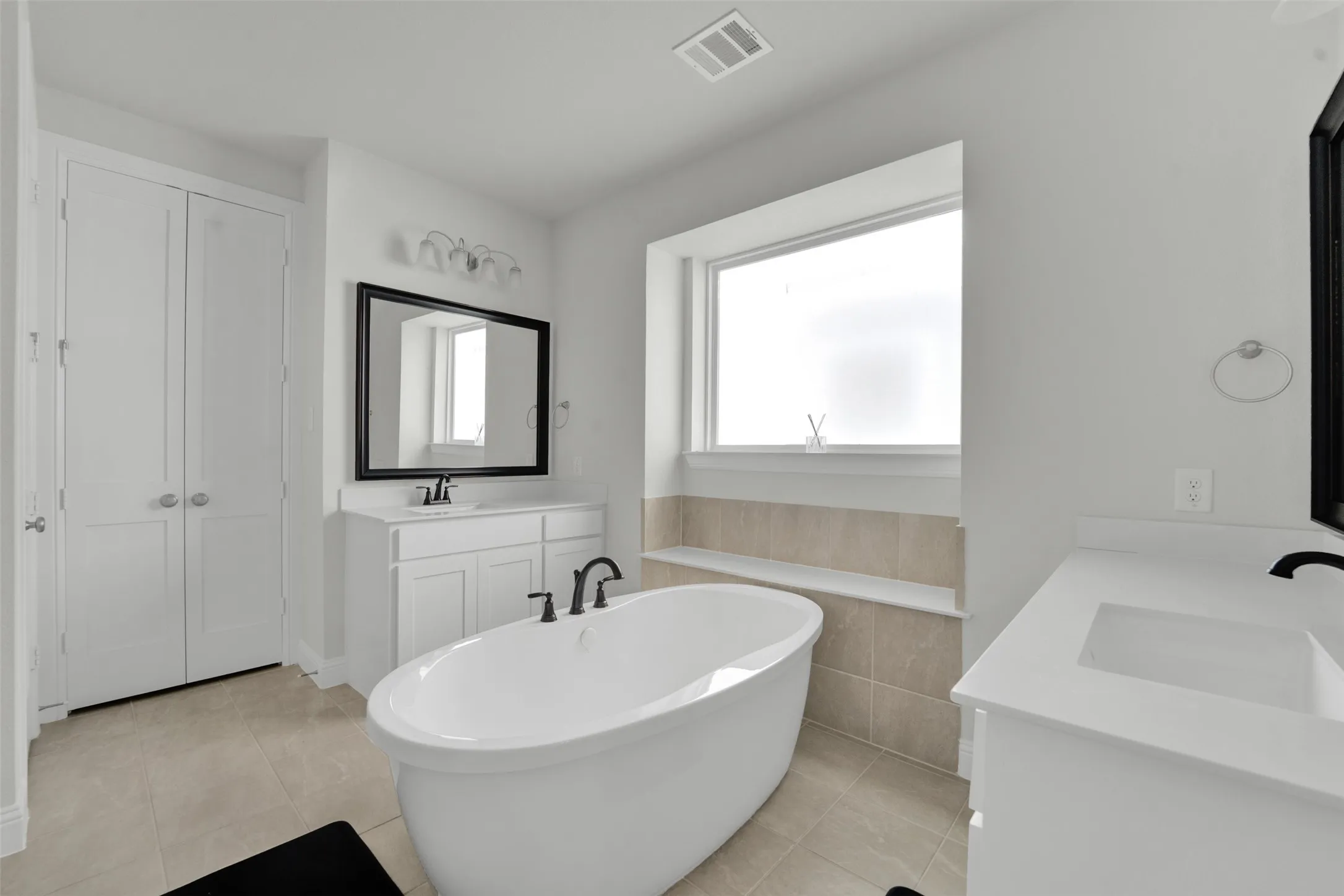 Freestanding soaking tub with matte black fixtures, dual vanity sinks with white countertops, and a window above a tiled bench