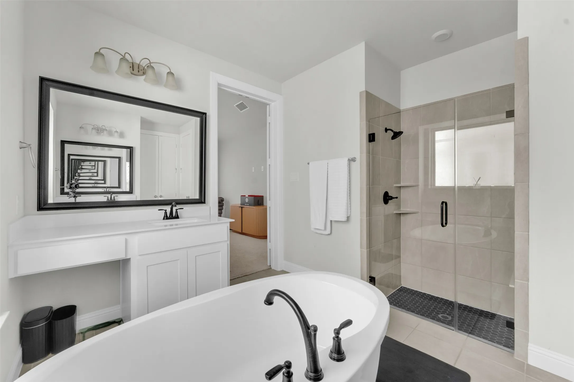 Contemporary bathroom featuring a freestanding soaking tub with matte black fixtures, a tiled walk-in shower with frameless glass enclosure, a spacious vanity with white cabinetry, a large framed mirror, and light-toned wall tile