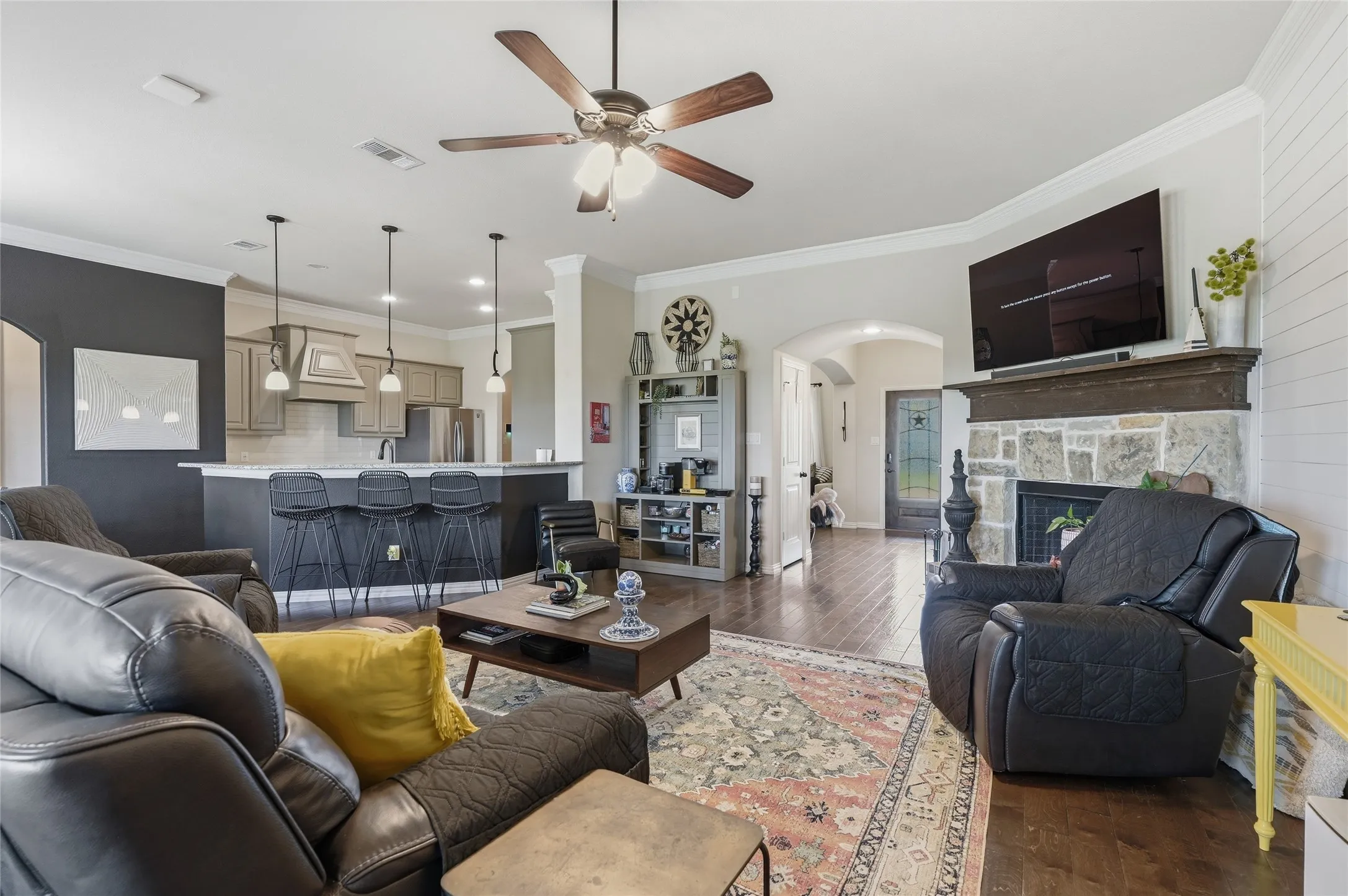Open concept living area featuring wood-finish flooring, a stone-front fireplace with a wood mantel, a ceiling fan, and crown molding