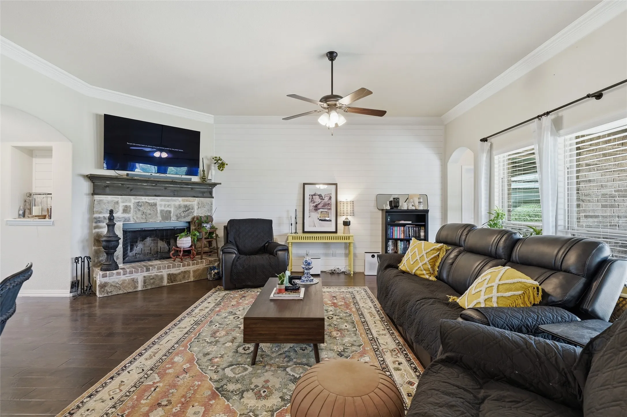 Living room featuring a stacked stone and brick fireplace with a wood mantel, dark wood-finish flooring, and crown molding
