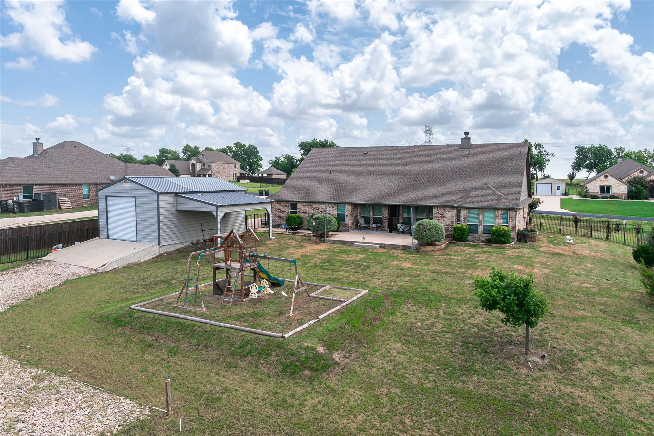 Expansive backyard featuring a detached garage with an attached carport, a large brick-faced residence with multiple windows, and a generous concrete patio