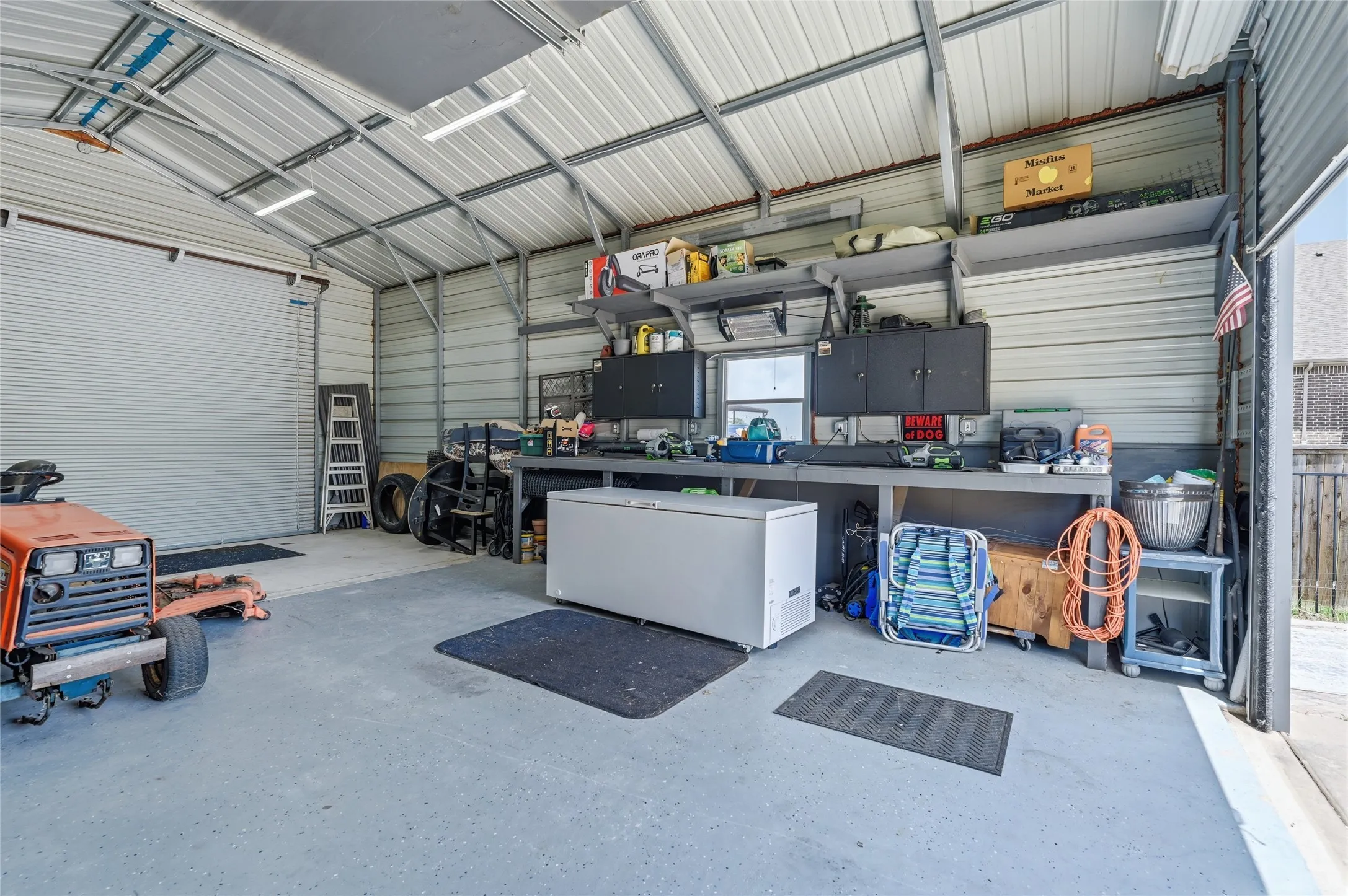 Metal construction carport featuring a roll-up garage door, concrete flooring, built-in workbench, and overhead storage shelving