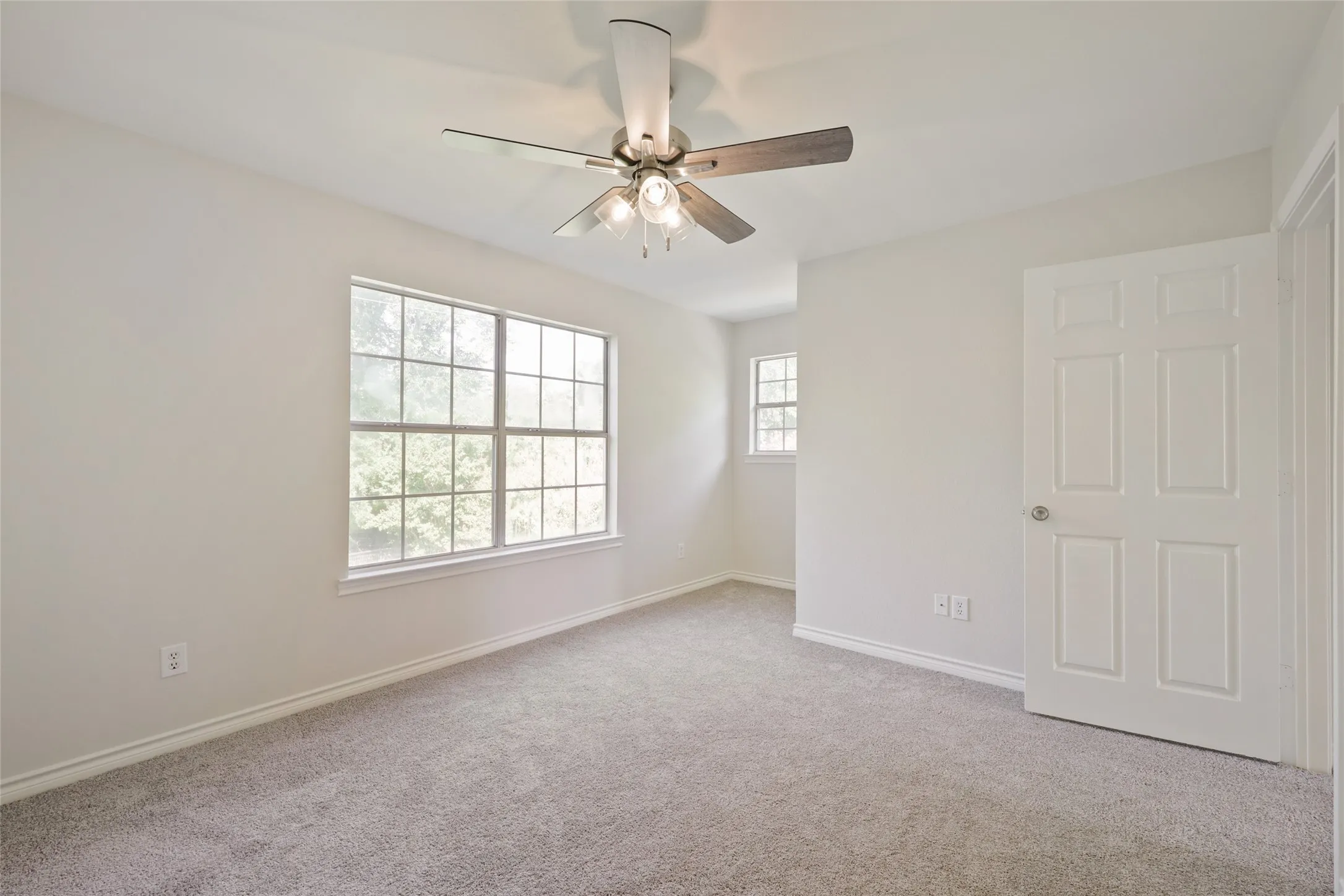 Carpeted room featuring a large window, a smaller window, a ceiling fan with integrated lighting, a paneled door, and white baseboards