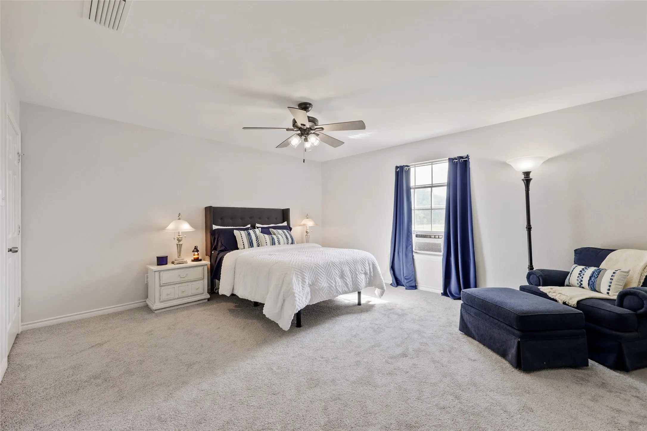 Spacious primary bedroom main home featuring light-colored carpeting, a ceiling fan with integrated lighting, and a window with blue drapes