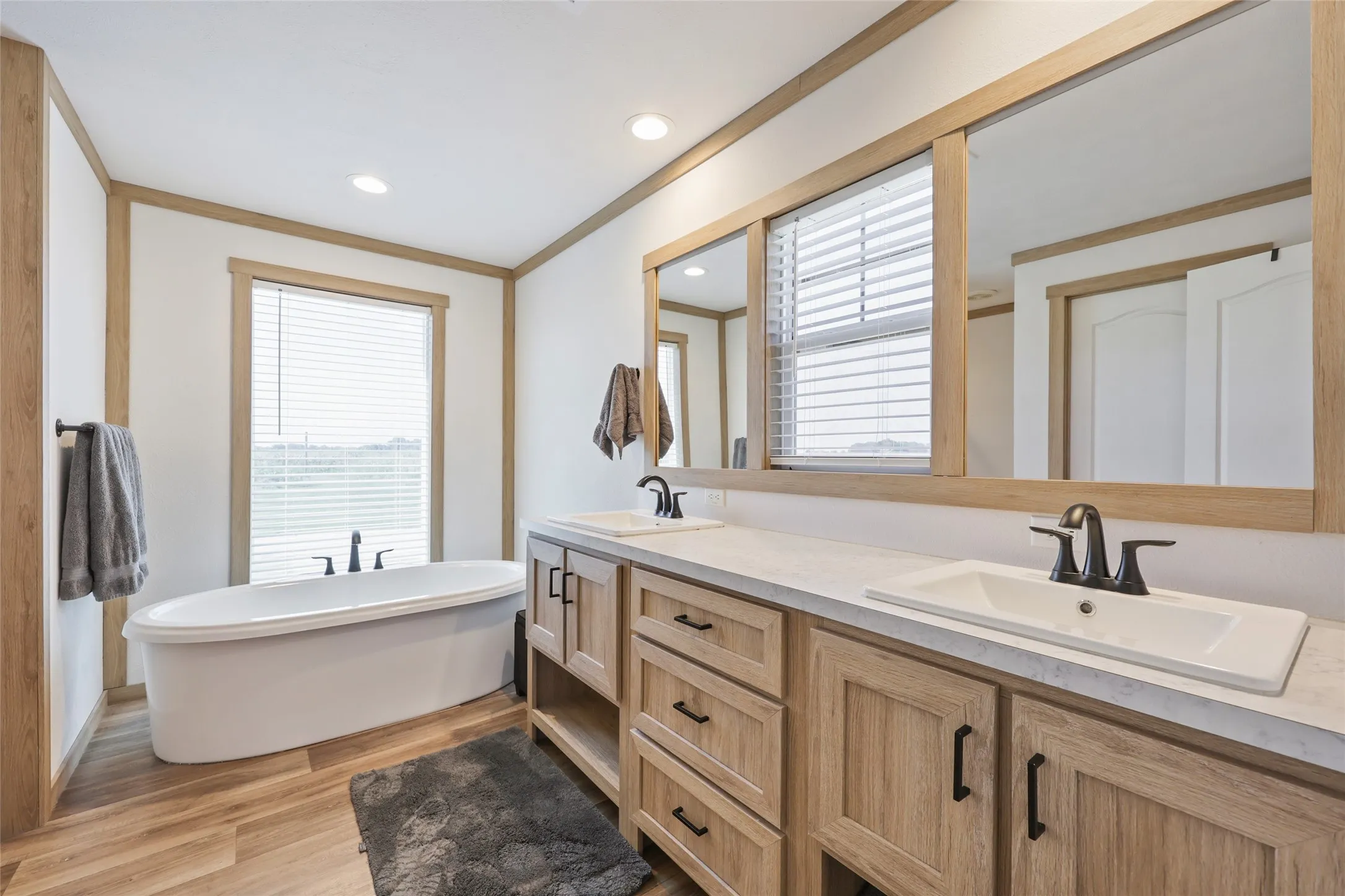 Freestanding soaking tub with a window located in 2nd home, dual vanity featuring light wood-finish cabinetry, integrated sinks, black hardware, and an expansive vanity mirror