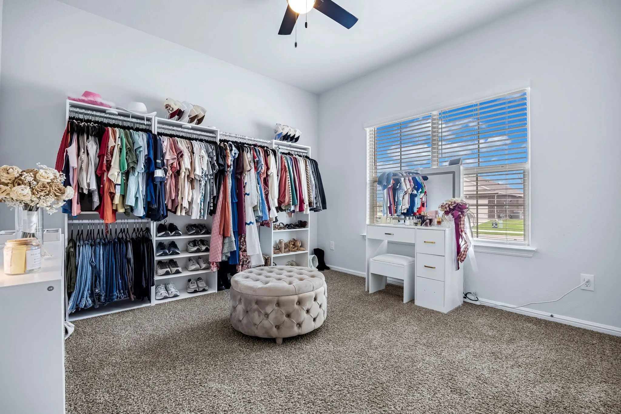 Spacious room featuring light gray walls, built-in open closet system with multiple hanging rods and shelving, a large window with white blinds, and carpeted flooring