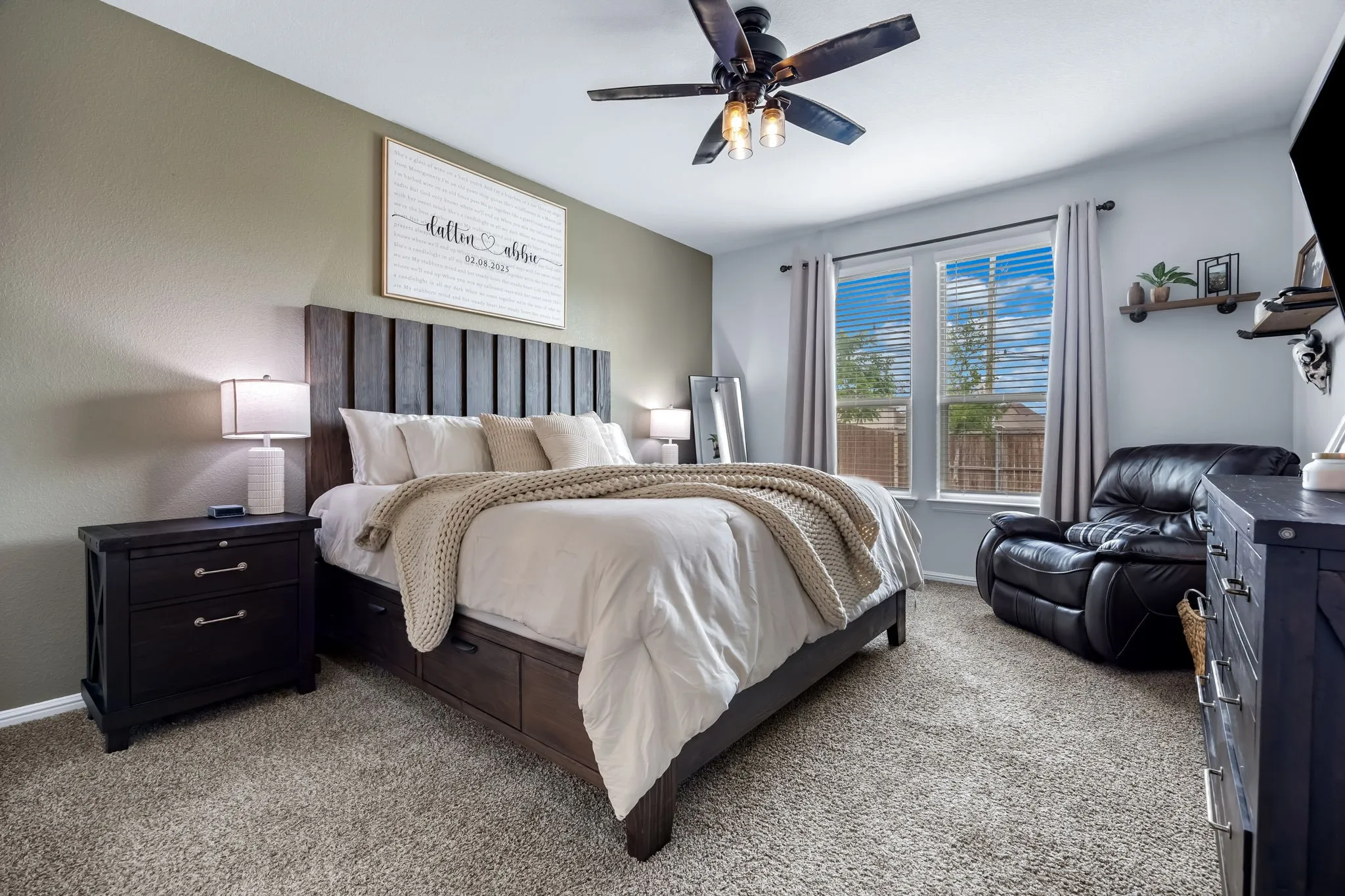 Carpeted bedroom featuring a ceiling fan with integrated lighting, two windows with blinds and drapes, and recessed shelving