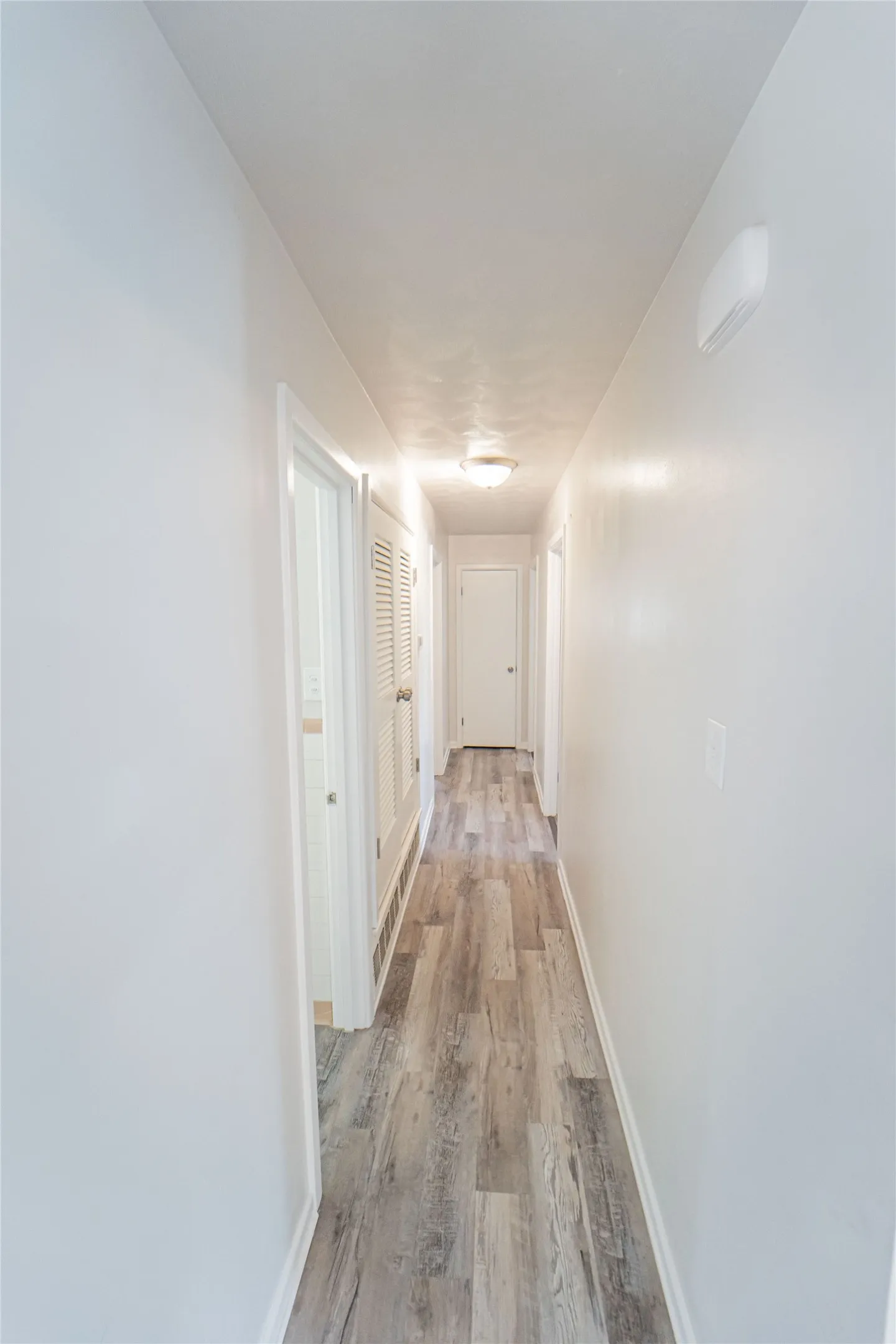 Hallway off of living area leading to guest bath and bedroom-featuring wood-finish flooring, white painted walls, and ceiling-mounted lighting fixtures