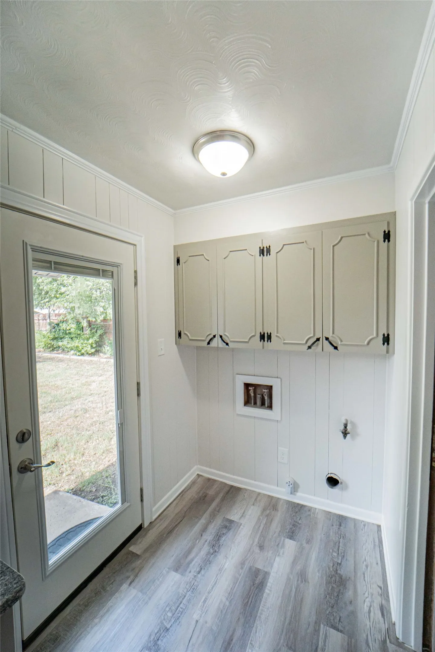 Light and bright-Laundry room located conveniently adjacent to kitchen and bedroom- featuring wood-finish flooring, white wainscoting, upper cabinetry with decorative paneling, and a full-light exterior door