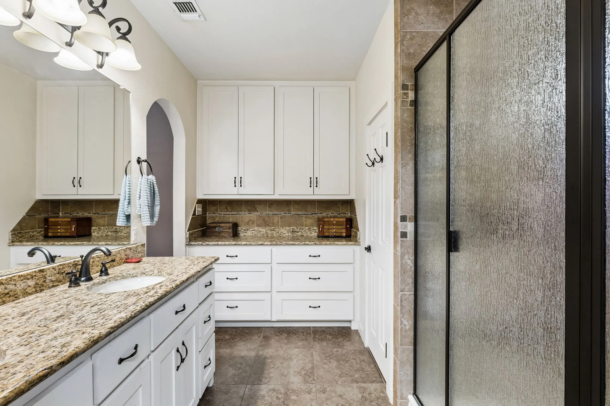 Bathroom featuring a double vanity with granite countertops, white cabinetry, dark bronze fixtures, a tiled backsplash, and a shower with a textured glass door