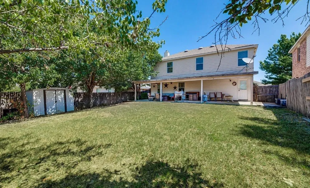 Expansive fenced backyard featuring a grassy lawn, mature trees, a covered patio, and a storage shed