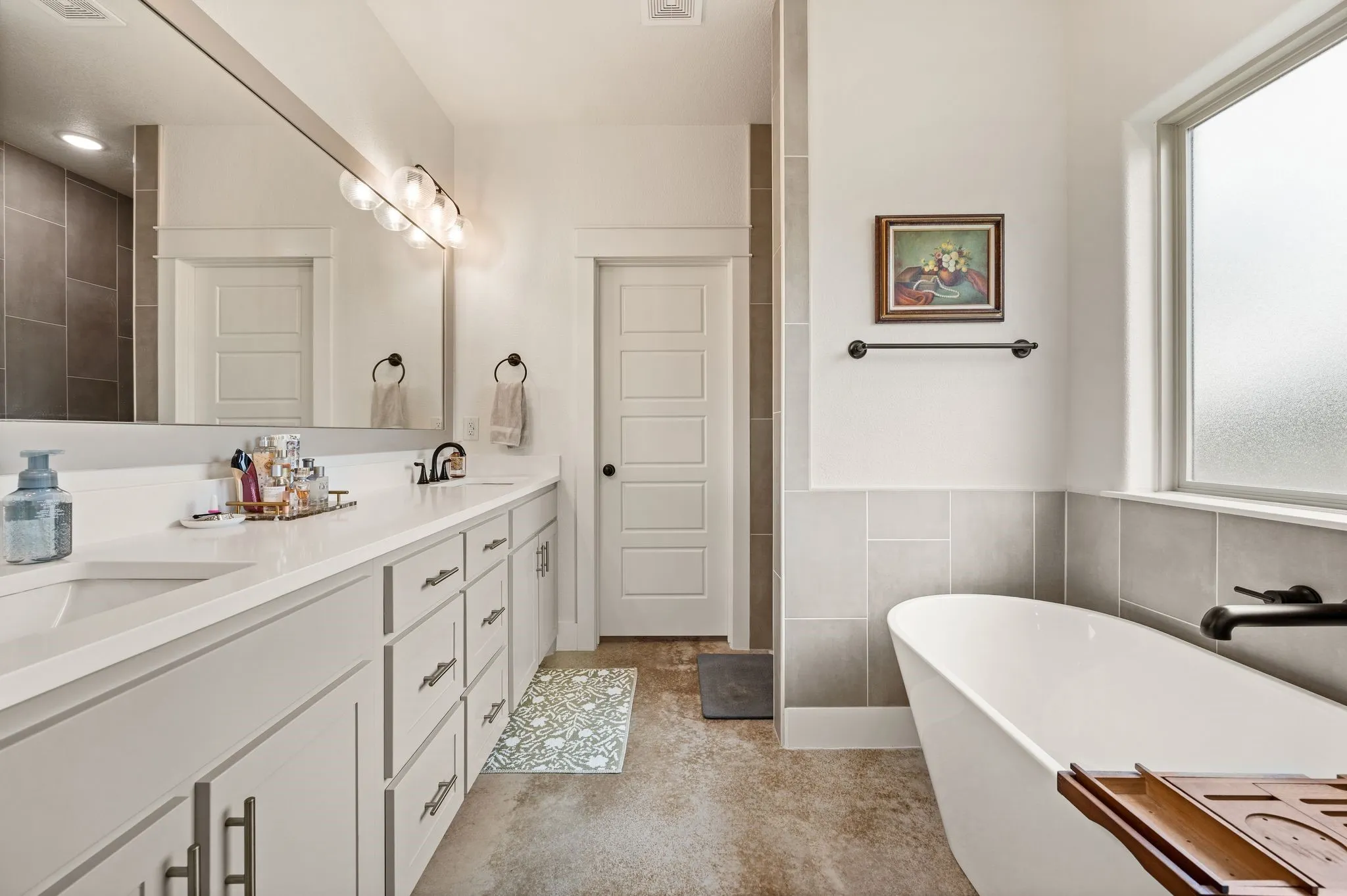Freestanding soaking tub with dark-finish fixtures, dual vanity with undermount sinks and light countertops, and a full-width mirror with sphere lighting