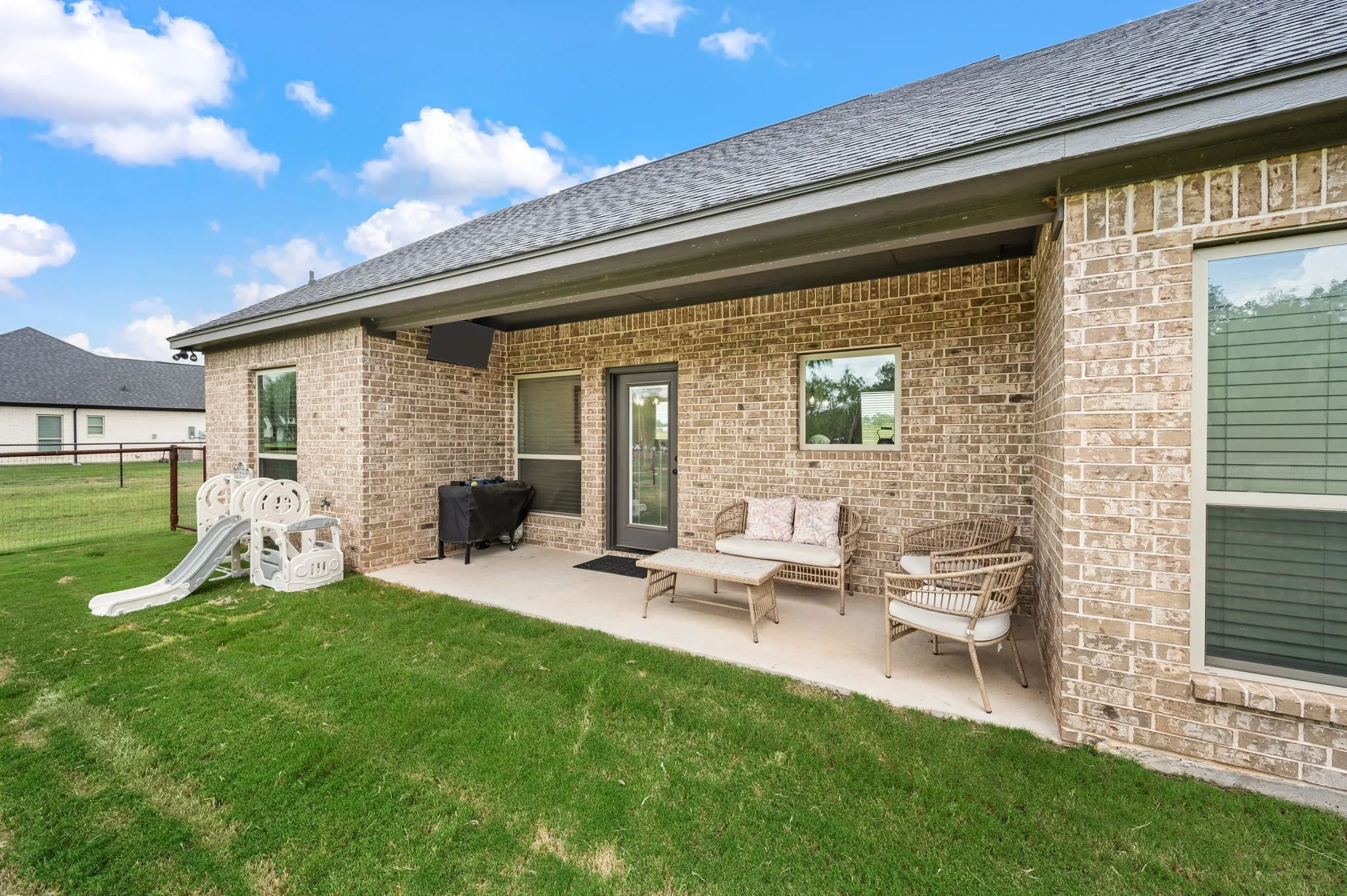 Expansive backyard featuring a covered concrete patio, brick exterior, dark-framed windows, and a fenced perimeter