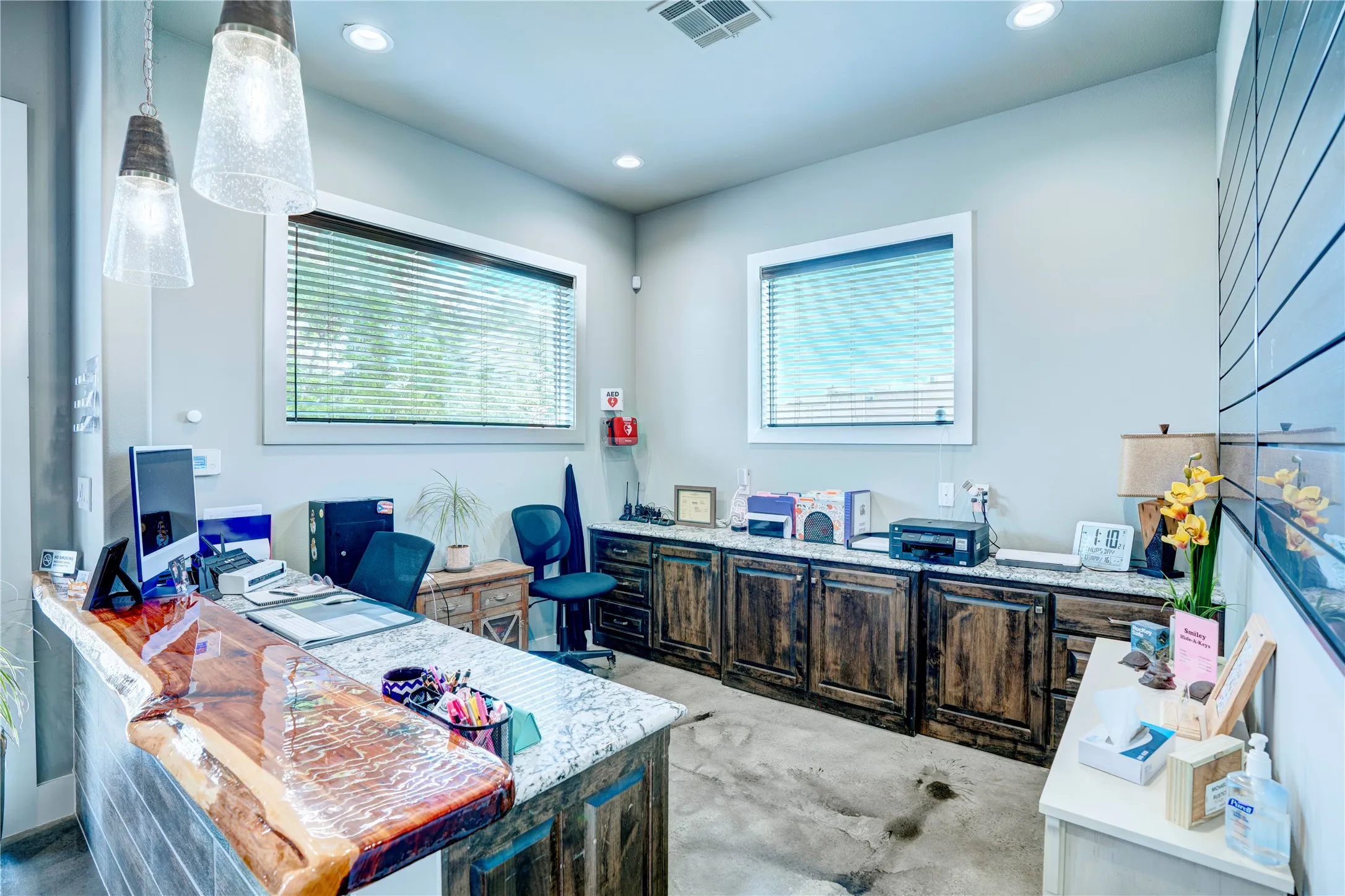 Reception area featuring a live-edge wood and stone reception desk, recessed ceiling lighting, two large windows with blinds, a built-in cabinetry run with stone countertops, and polished concrete flooring