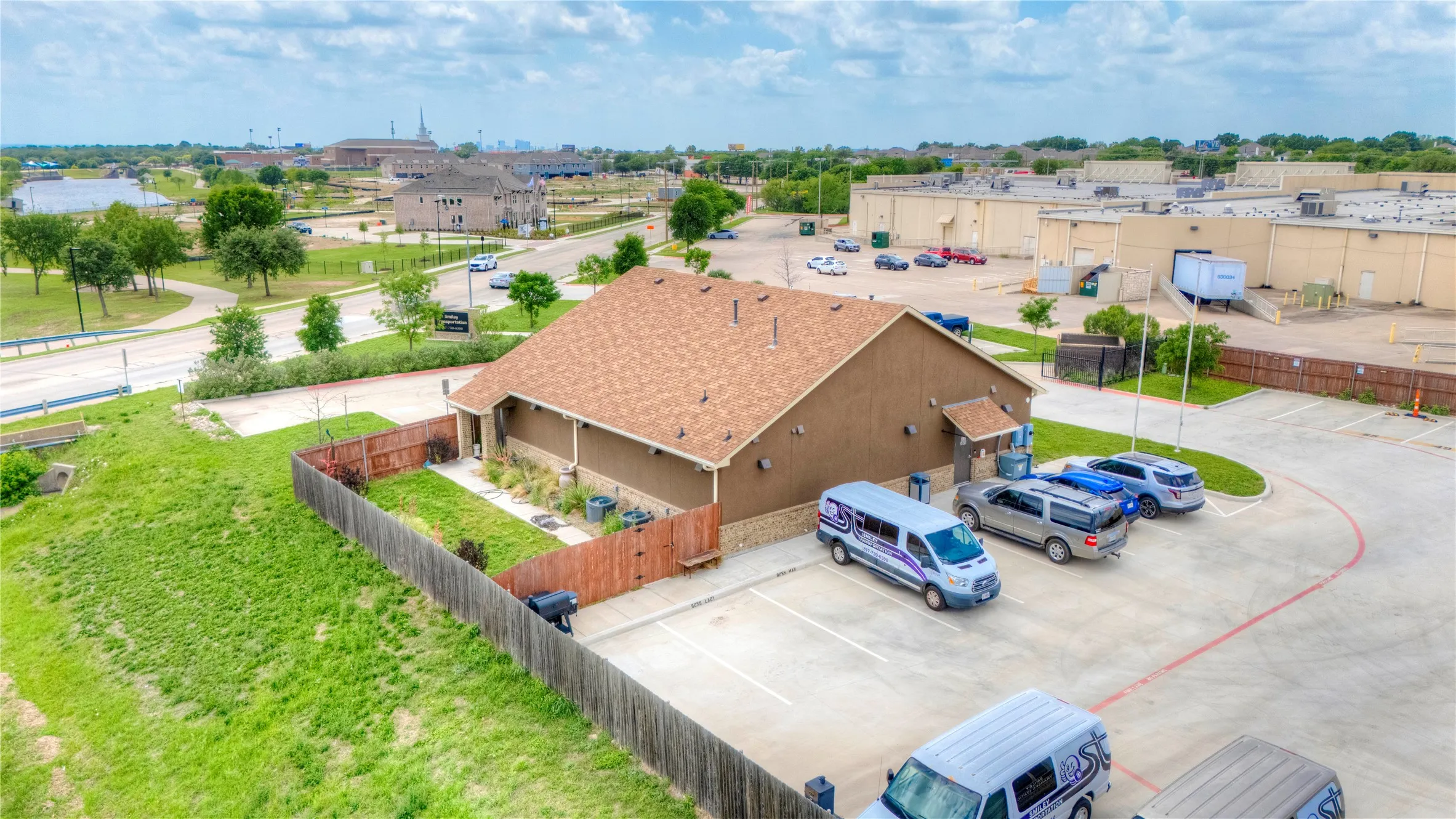 Low-rise building with a brown composite shingle roof and stucco exterior, featuring a private fenced yard, a paved parking area, and mature landscaping