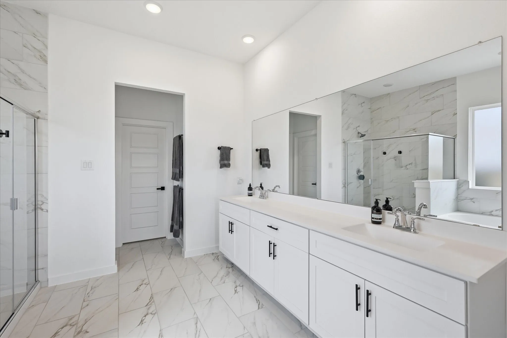 Spacious bathroom featuring a dual vanity with white cabinetry and chrome fixtures, a large frameless mirror, and a glass-enclosed shower with white marble-style tile
