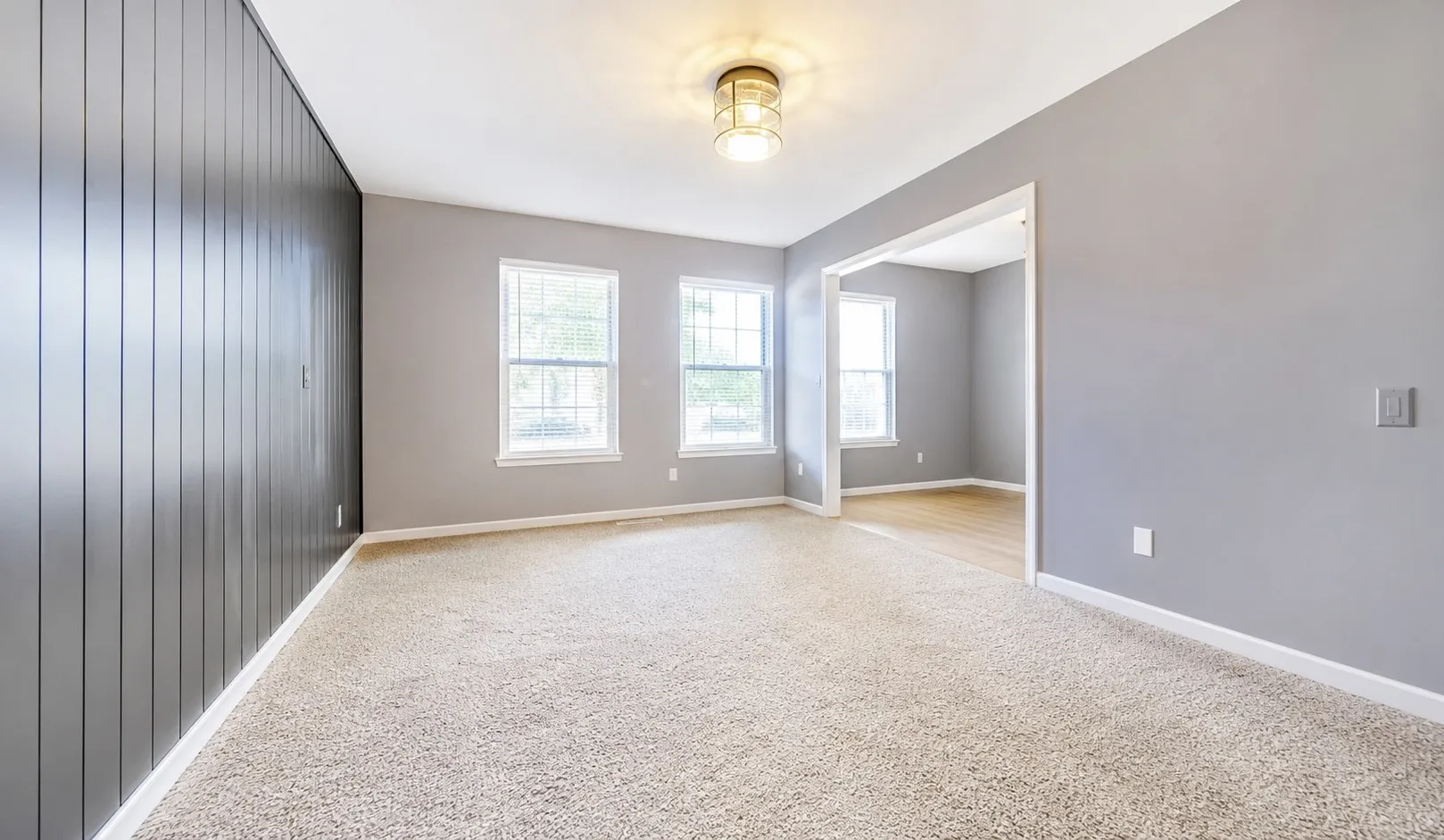 Carpeted room featuring a dark accent wall with vertical paneling, three windows, a ceiling-mounted light fixture, and an open doorway to an adjoining room with wood-finish flooring