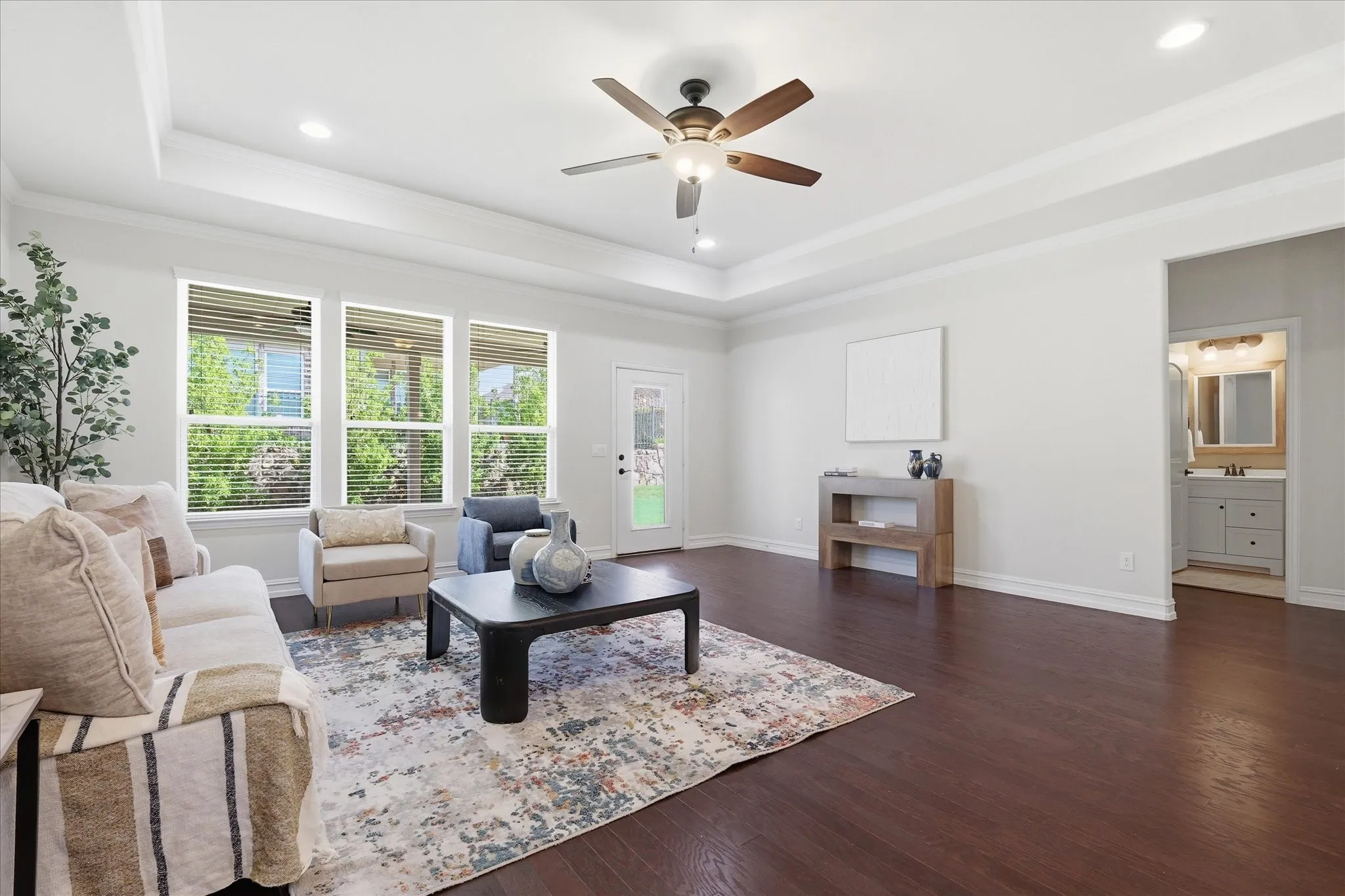 Spacious living area featuring rich wood-finish flooring, a tray ceiling with recessed lighting and a ceiling fan, and three large windows with blinds