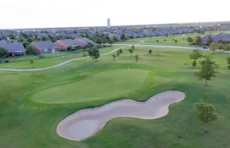 Manicured golf course green with a sand trap, surrounded by lush grass and mature trees