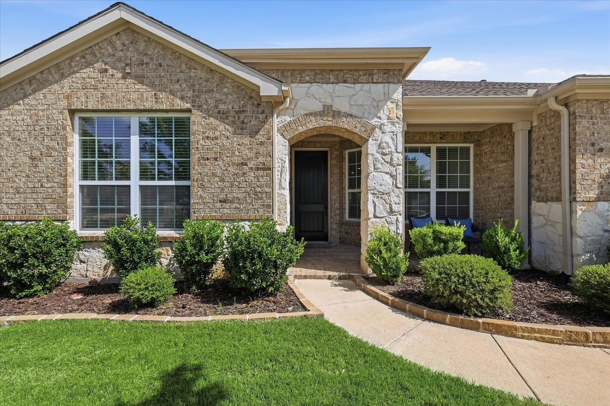 Brick and stone facade featuring an arched entry, multiple large windows with grids, a prominent front walkway, and structured landscaping