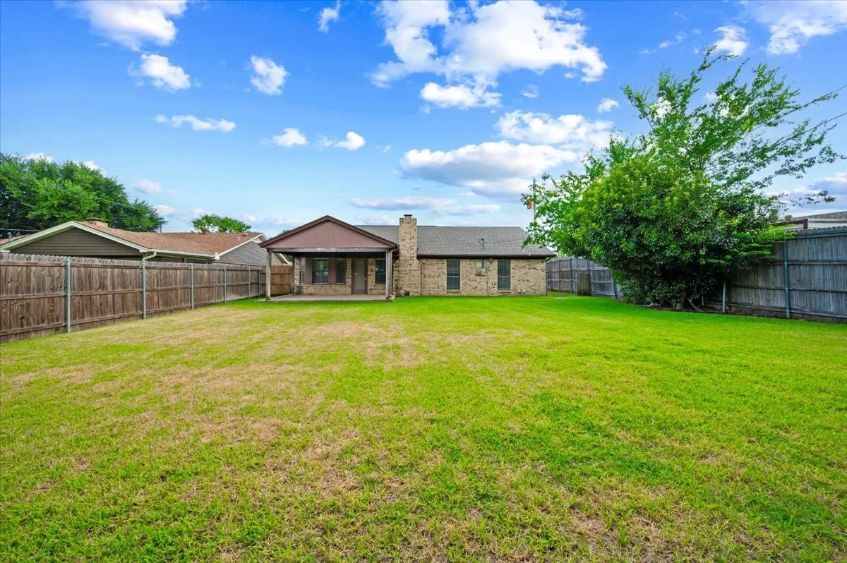 Expansive backyard featuring a large green lawn, full privacy fencing, a covered patio, and mature trees providing natural shade