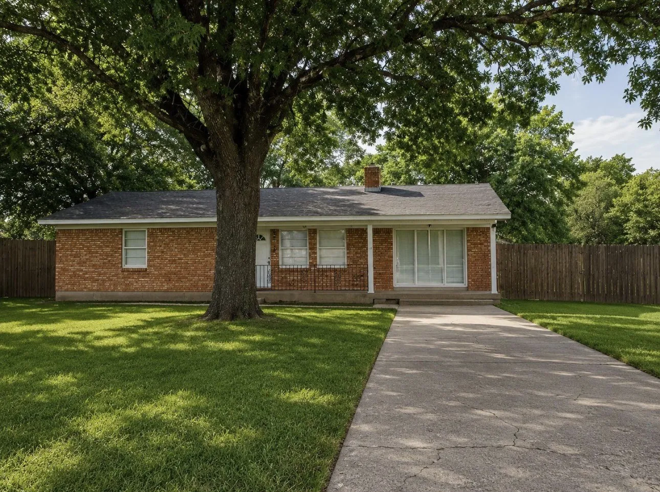 Brick exterior ranch-style home featuring a covered front porch with white railing, a large mature tree, a paved driveway, and a fenced perimeter