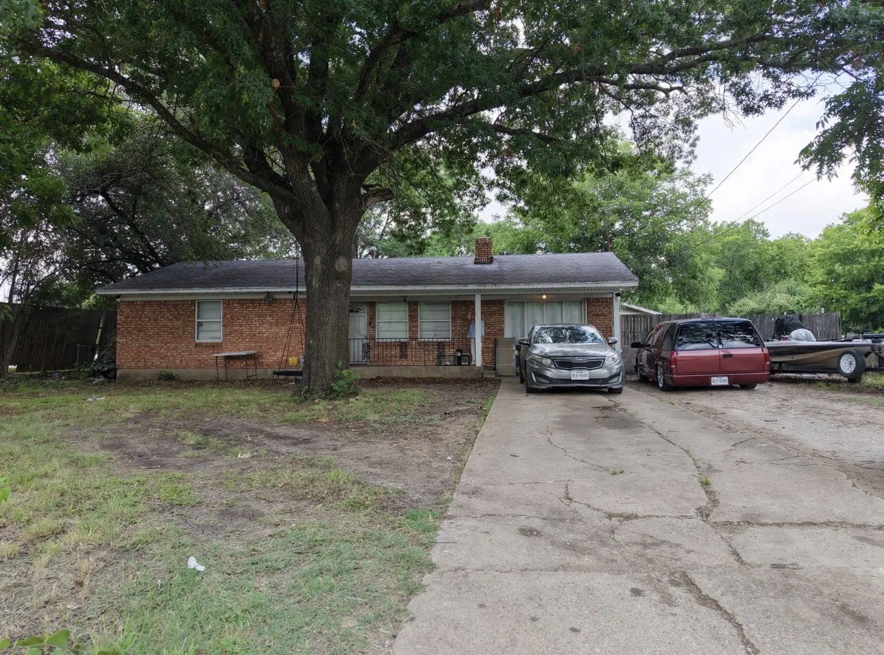 Brick exterior home featuring a covered front porch, a mature shade tree, a concrete driveway, and a fenced perimeter