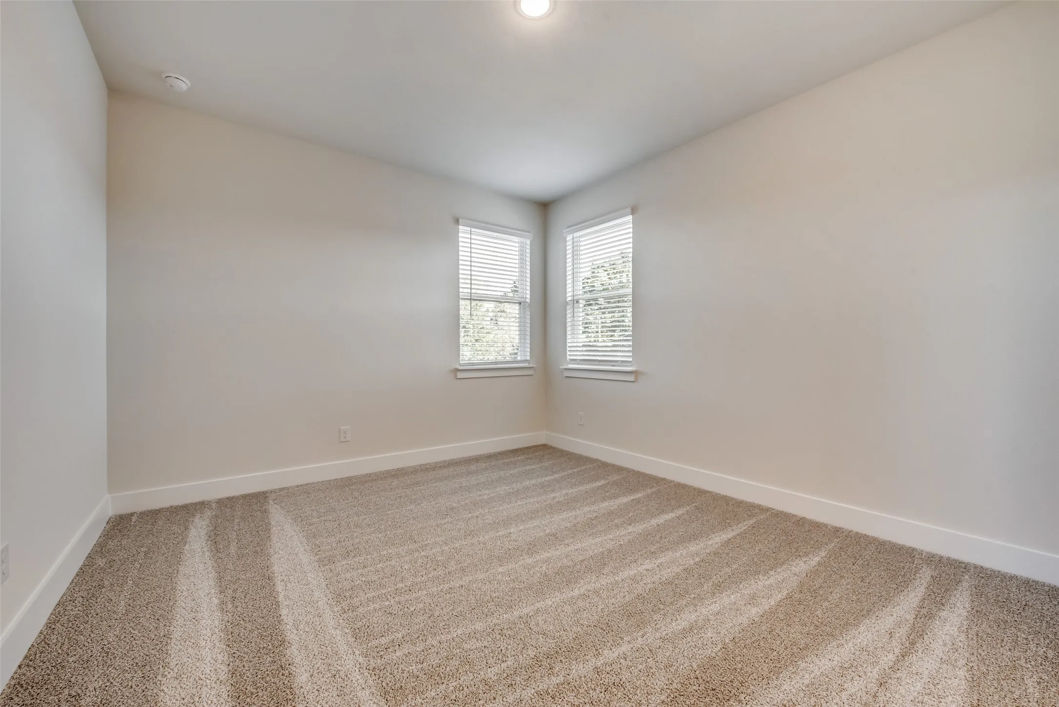 Corner windows with white blinds, neutral wall paint, white baseboards, and light-colored carpet flooring