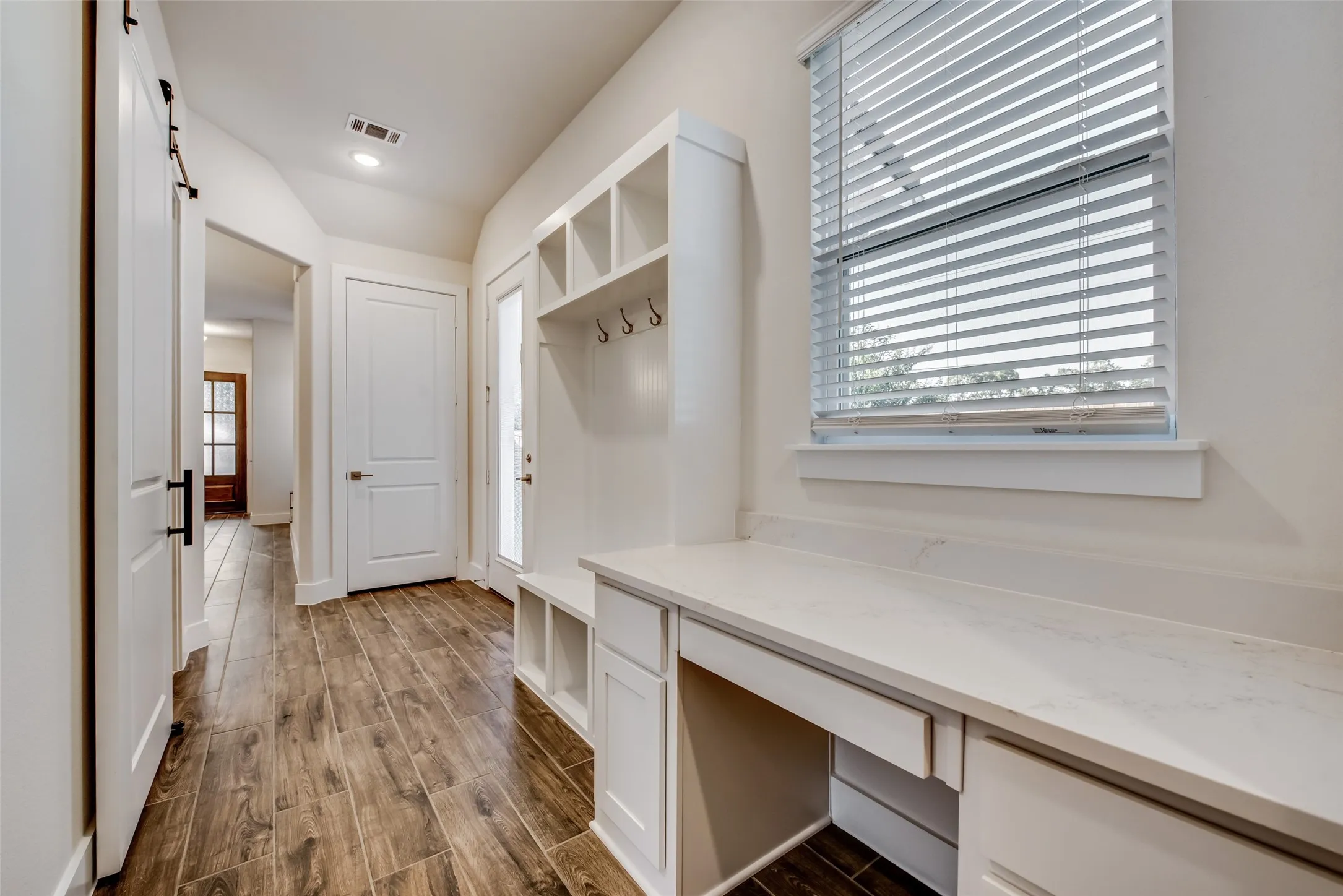 Entryway featuring wood-finish flooring, a built-in organizational unit with hooks and cubbies, a built-in desk with a light-toned countertop, a window with blinds, and a barn door